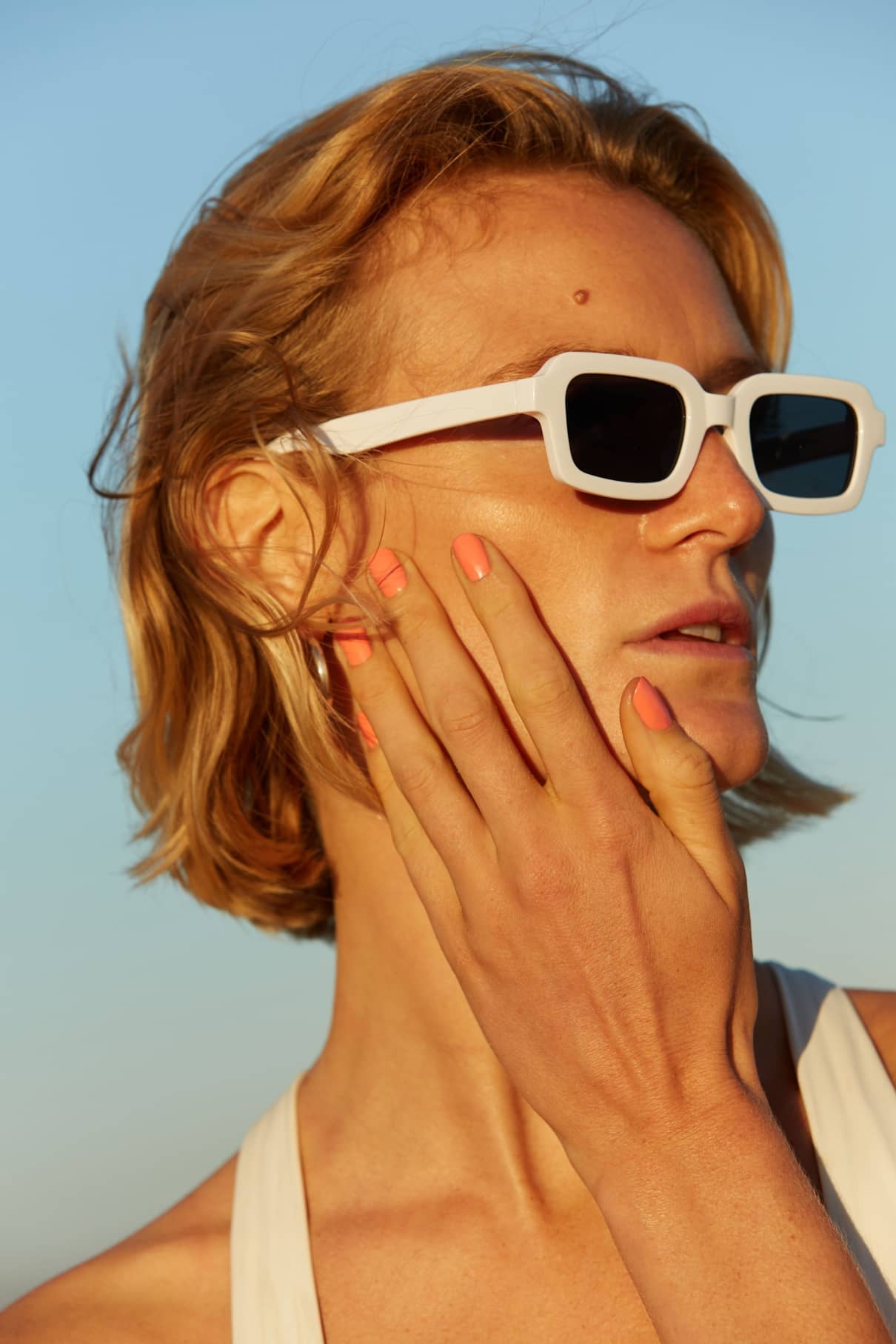 Eine Person mit kurzen blonden Haaren trägt eine weiße Sonnenbrille, berührt ihr Gesicht und präsentiert gitti no. 010 pfirsichfarbenen Nagellack in einem zarten Korallenton; sie befindet sich im Freien im Sonnenlicht.