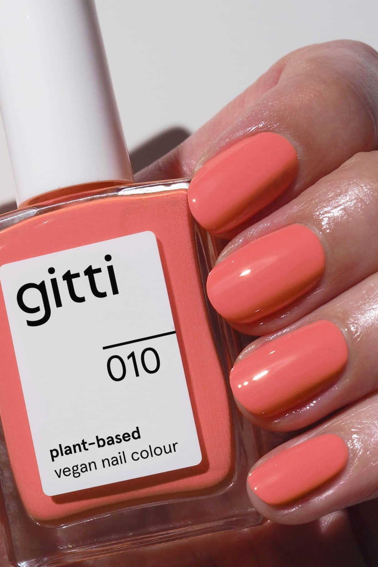 Eine Hand mit sauber manikürten Nägeln, die in einem leuchtenden Korallenton lackiert sind, hält eine Flasche mit gitti no. 010 pflanzlichen veganen Nagellack, der perfekt zur Nagelfarbe passt.