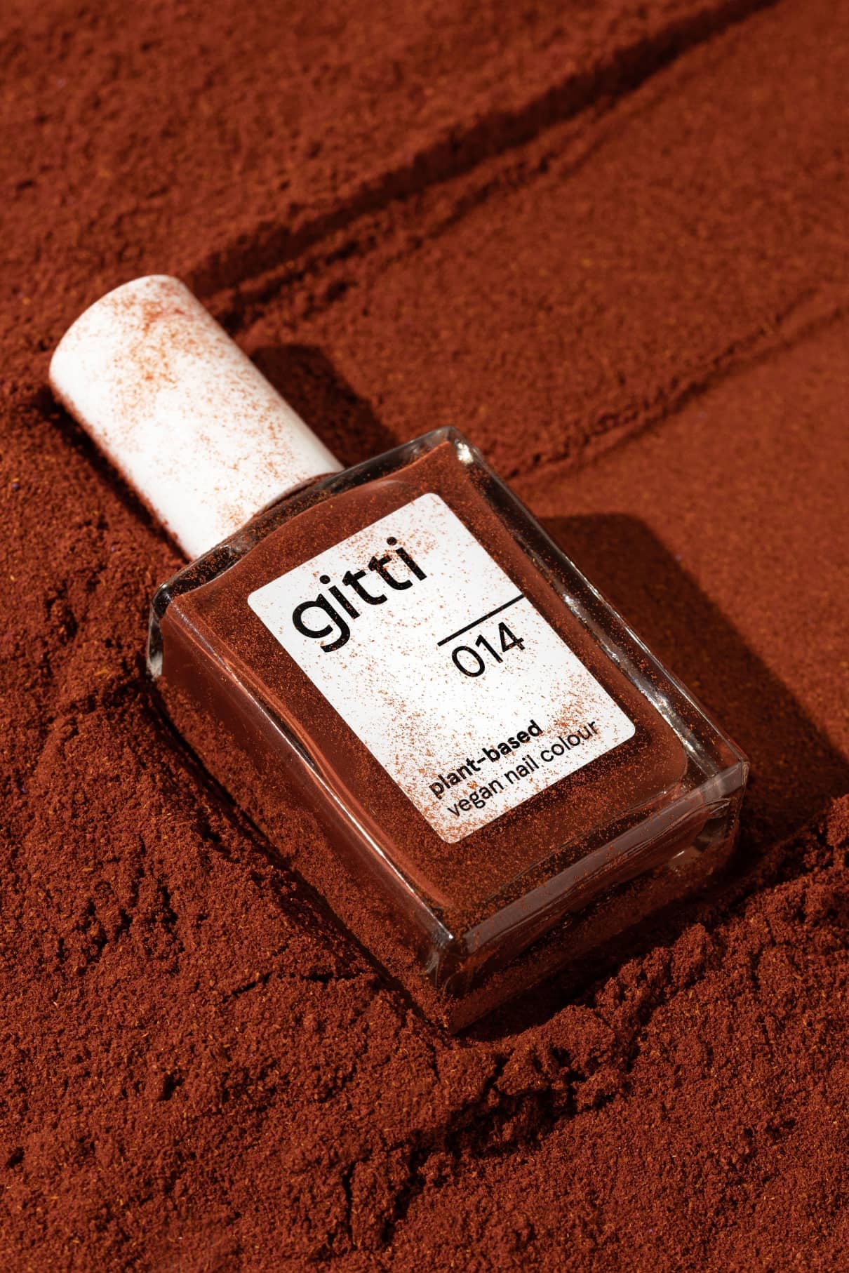 Eine Flasche mit gitti no. 014 pflanzliche, vegane Nagelfarbe ruht auf einer mit warmem, rötlich-braunem Puder bestäubten Oberfläche.