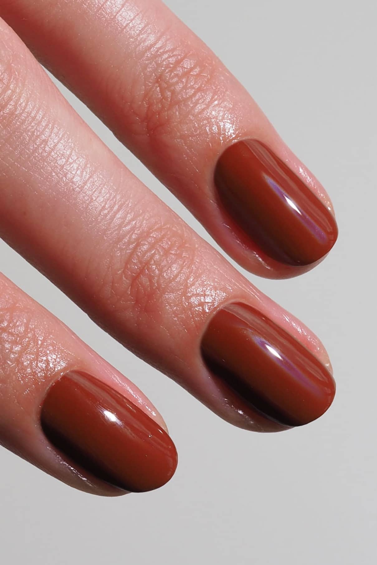 Eine Nahaufnahme von drei Fingern mit kurzen, ovalen Nägeln, die mit glänzendem Gitti No. 014 Nagellack, einem warmen Braunton, vor einem unifarbenen Hintergrund.