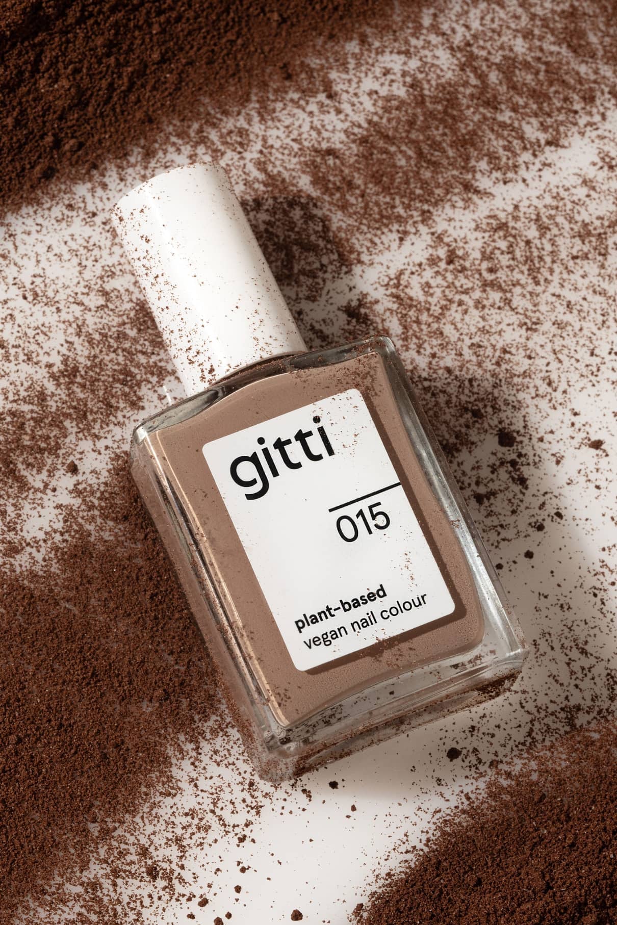 Ein Fläschchen mit gitti no. 015 pflanzlicher veganer Nagellack in cremigem Beige ruht auf einer weißen, mit braunem Puder bestäubten Fläche.