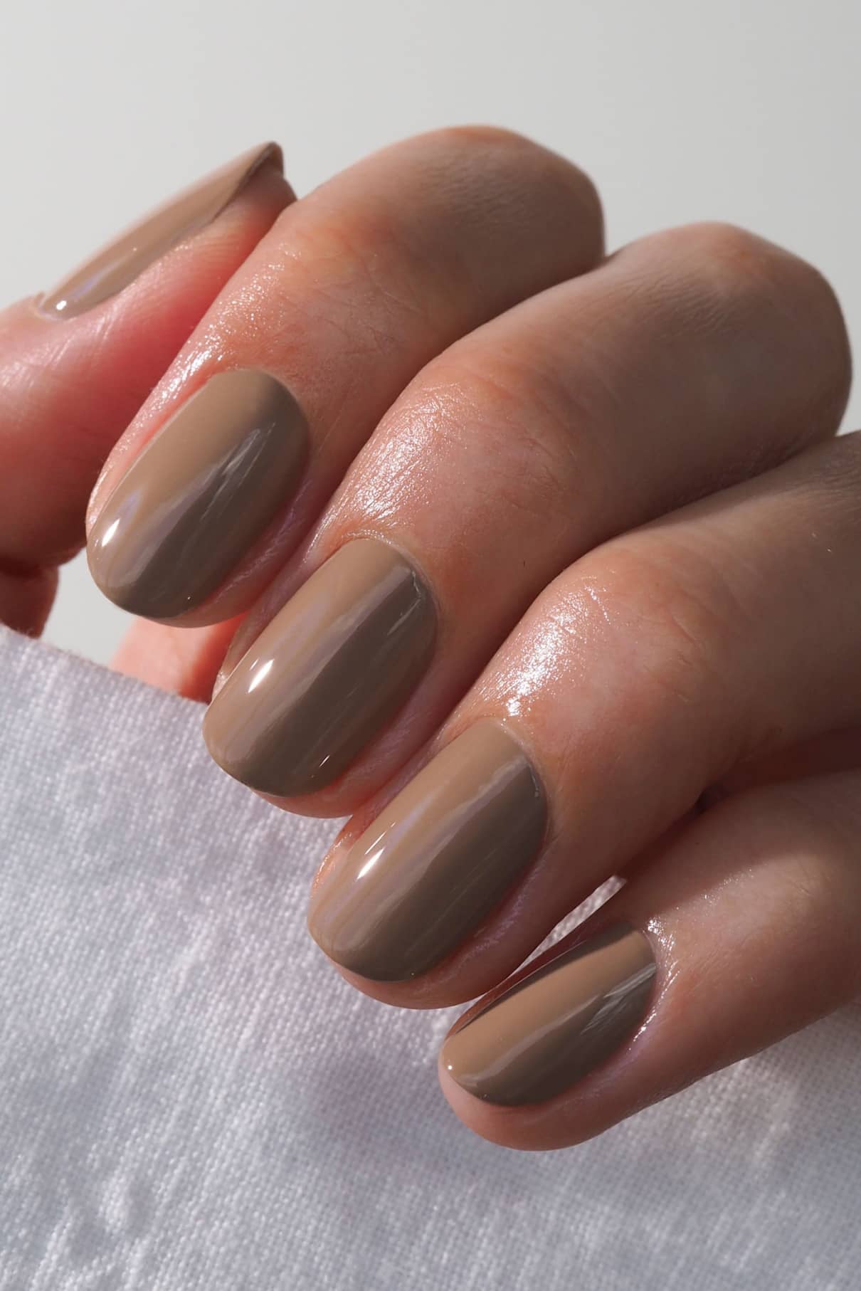 Eine Hand mit kurzen, sauber manikürten Nägeln, die in glänzendem dunklem Beige (gitti Nr. 015) lackiert sind, hält einen weißen, strukturierten Stoff.