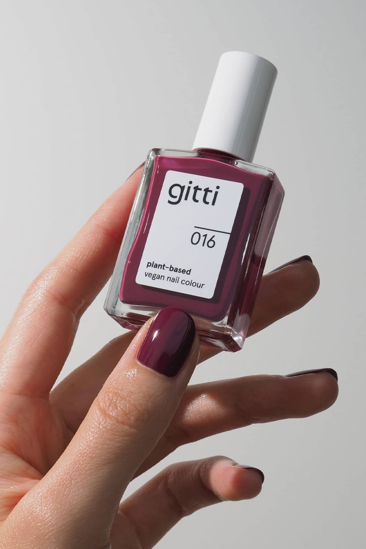 Eine Hand mit lackierten Nägeln hält eine Flasche von gitti no. 016, einer pflanzlichen, veganen Nagelfarbe in Beerenrot, einem tiefen Lila, das von trendigen Lippenstiften inspiriert ist.