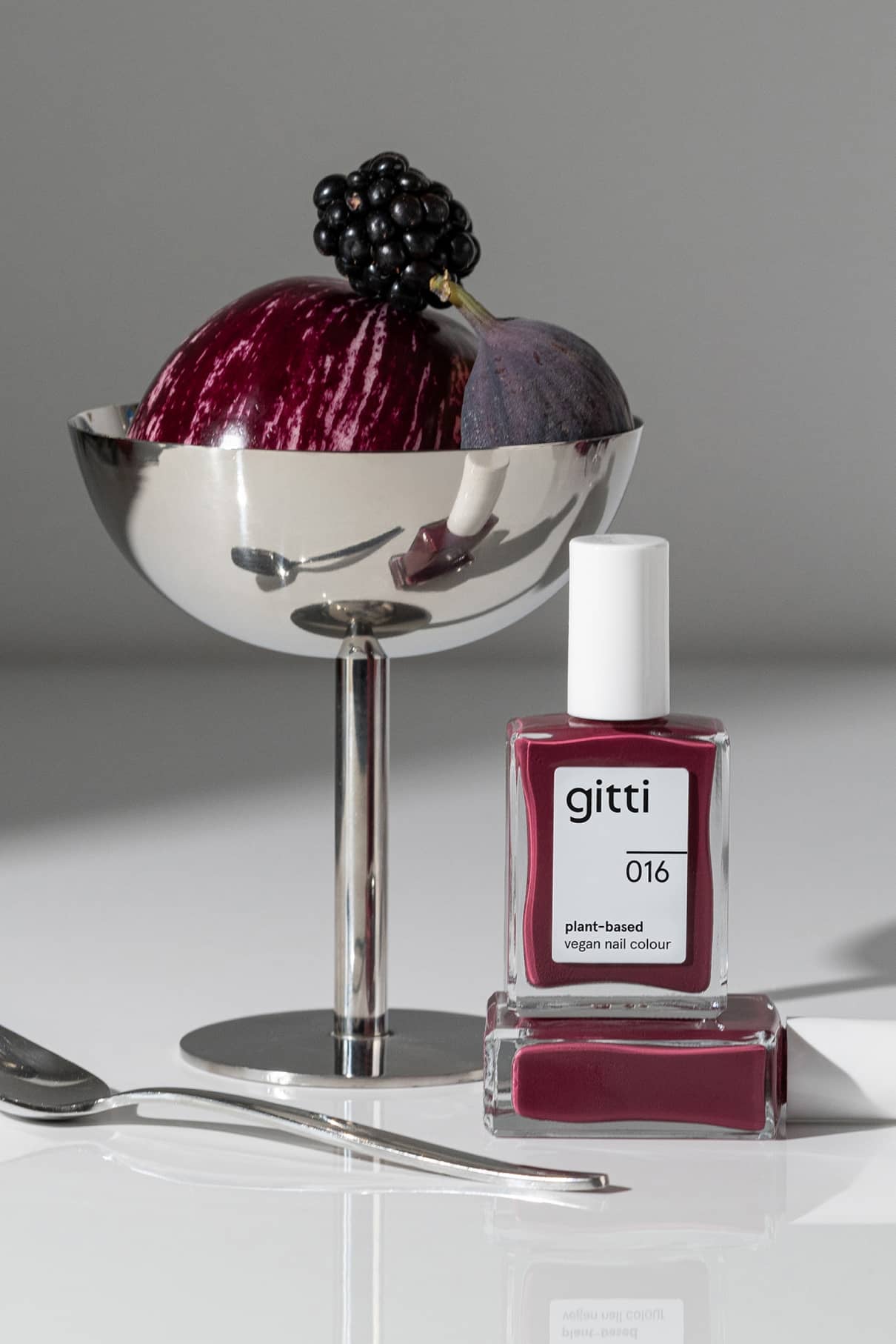 Eine Flasche gitti no. 016 pflanzlicher Nagellack in Beerenrot steht neben einer Metallschale mit einer halbierten Feige und einer Brombeere, die von einem Löffel begleitet wird - und den Charme des dunkelroten Farbtons eines trendigen Lippenstifts aufgreift.