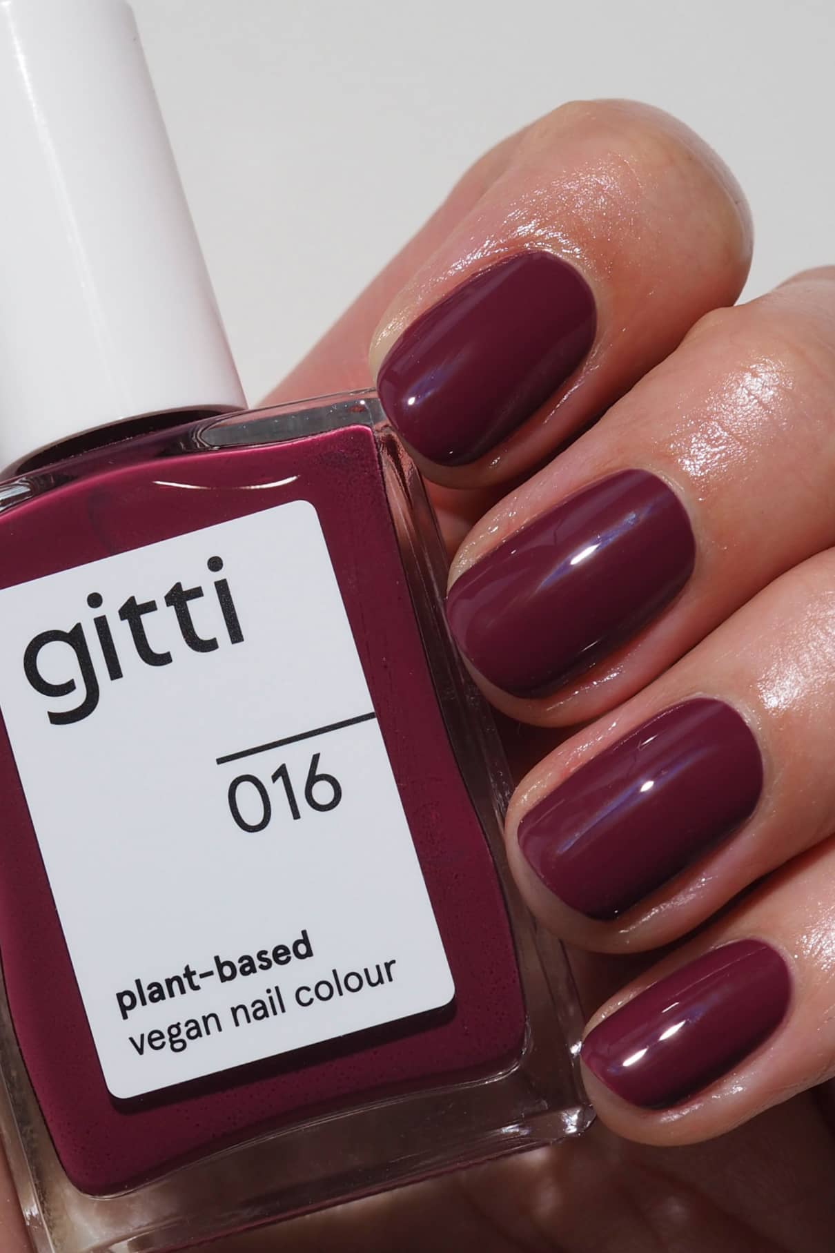 Eine Hand mit kurzen, gepflegten, in Beerenrot lackierten Nägeln hält eine Flasche gitti no. 016 pflanzlichen, veganen Nagellack, der perfekt zu deinem Lieblingslippenstift passt.