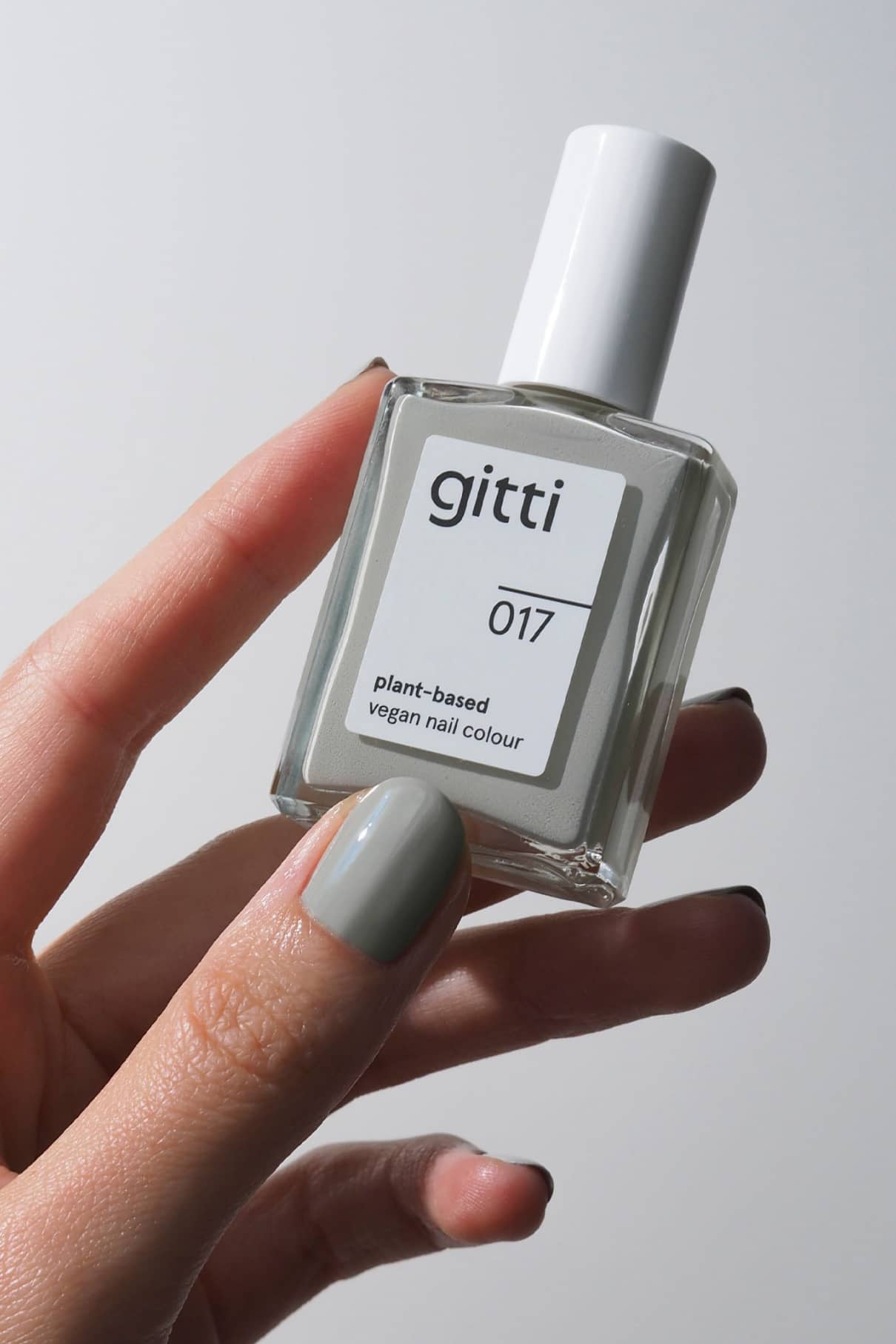 Eine Hand mit grau lackierten Nägeln hält eine Flasche von gitti no. 017, eine pflanzliche, vegane Nagelfarbe in hellem Grau - ein idealer Winterton - vor einem schlichten Hintergrund.