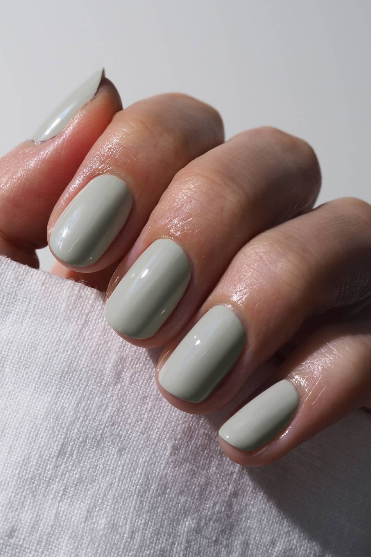 Eine Hand mit gepflegten Nägeln in gitti no. 017, einem hellen Grau mit salbeigrünem Unterton, hält einen weißen Stoff - die winterliche Version moderner Eleganz.