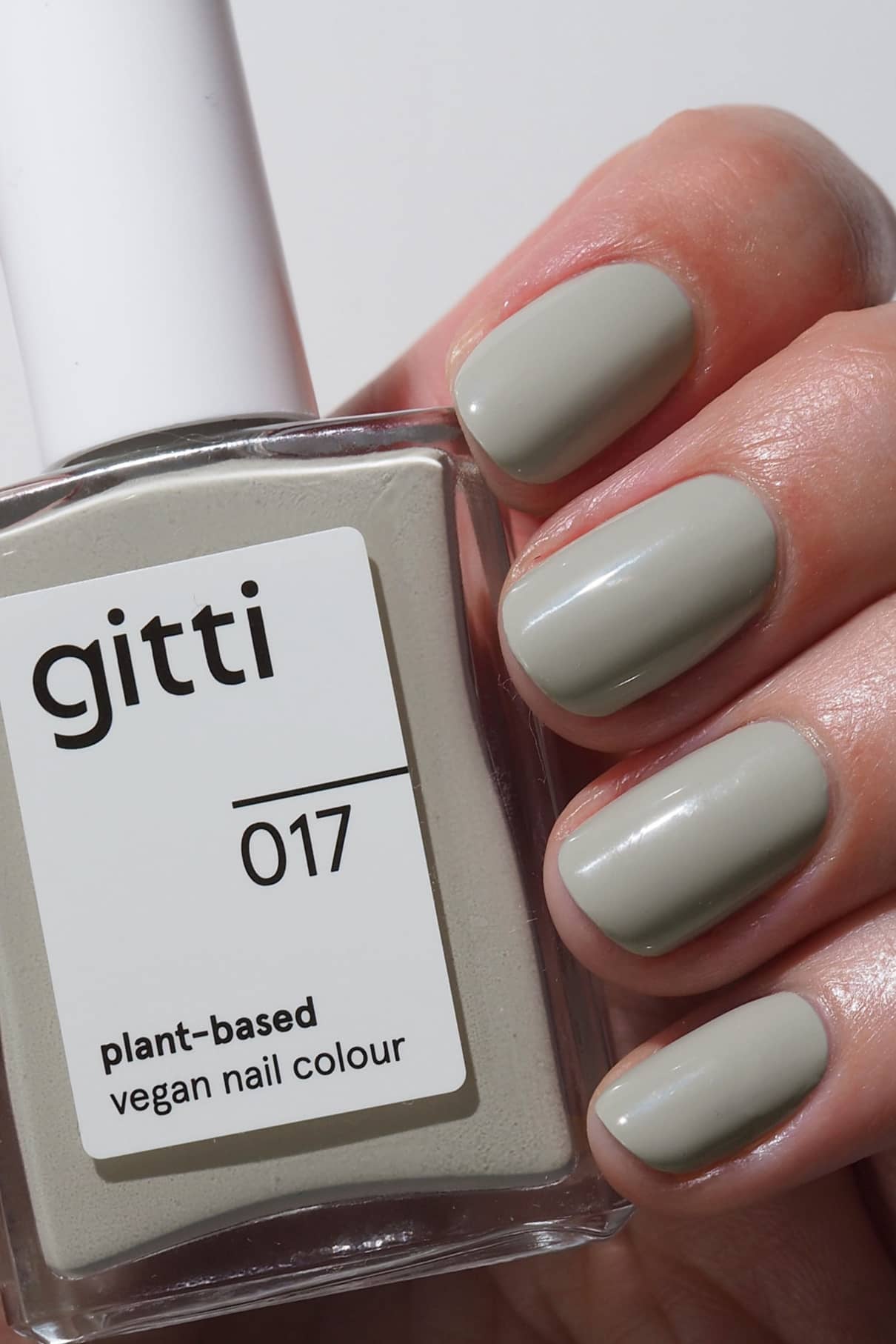 Eine Hand mit kurzen, sauber manikürten Nägeln, die in Salbeigrün lackiert sind - einem winterlichen, hellgrauen Grünton - hält eine Flasche mit der Aufschrift gitti no. 017, eine vegane Nagelfarbe auf Pflanzenbasis.