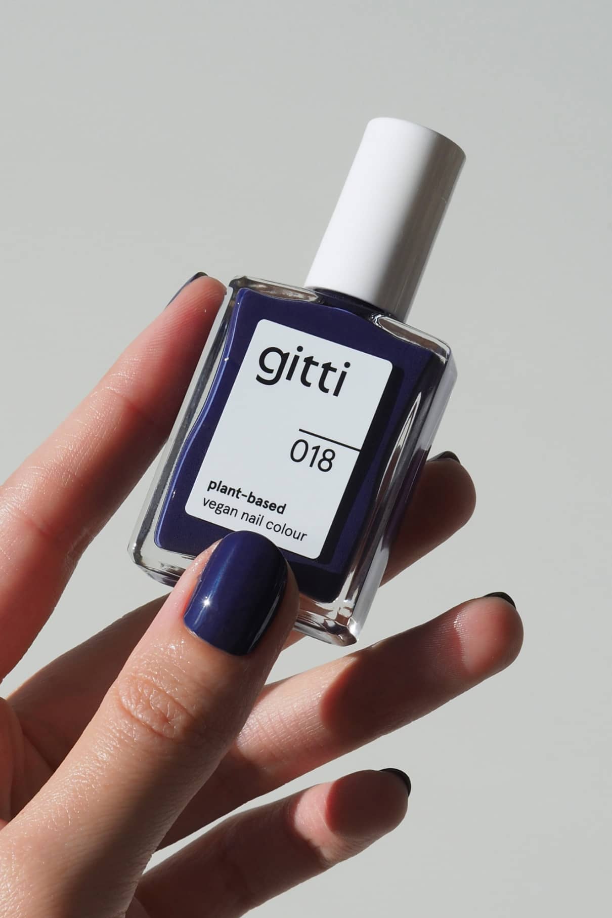 Eine Hand mit lackierten Nägeln hält eine Flasche von gitti no. 018, einer pflanzlichen, veganen Nagelfarbe in einer Mischung aus Tiefblau und Violett.
