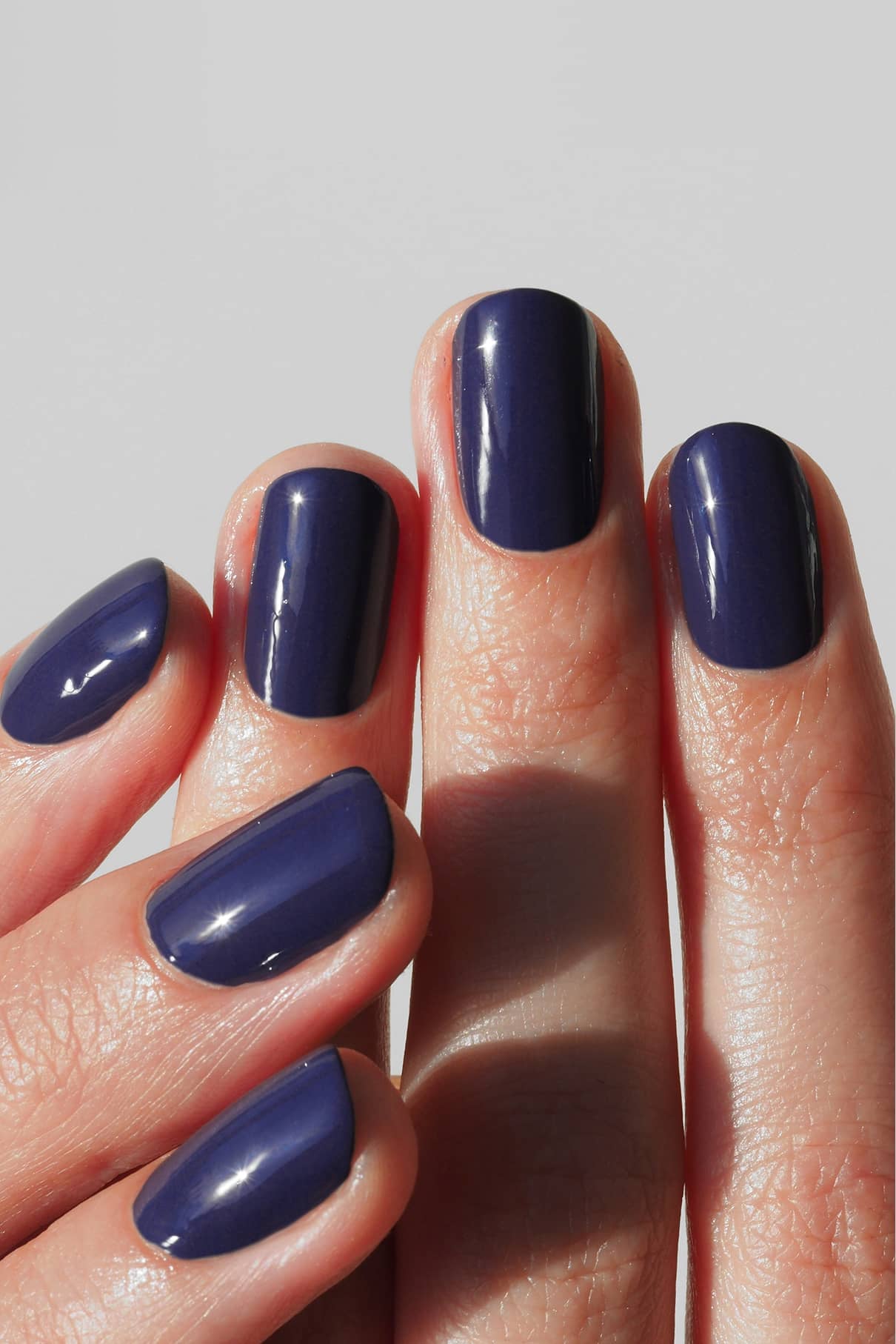 Eine Hand mit sauber manikürten Nägeln, lackiert mit gitti no. 018, einem glänzenden, tiefblauen Lack mit einem Hauch von Violett, vor einem hellen Hintergrund.
