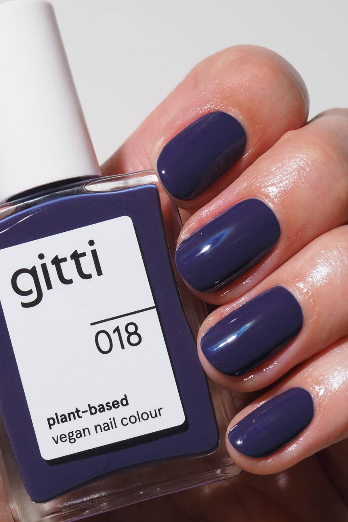 Eine Hand mit sauber manikürten Nägeln, die in einer Mischung aus tiefem Blau und Violett lackiert sind, hält eine Flasche gitti no. 018, einem veganen Nagellack auf Pflanzenbasis, in der gleichen kräftigen Nuance.