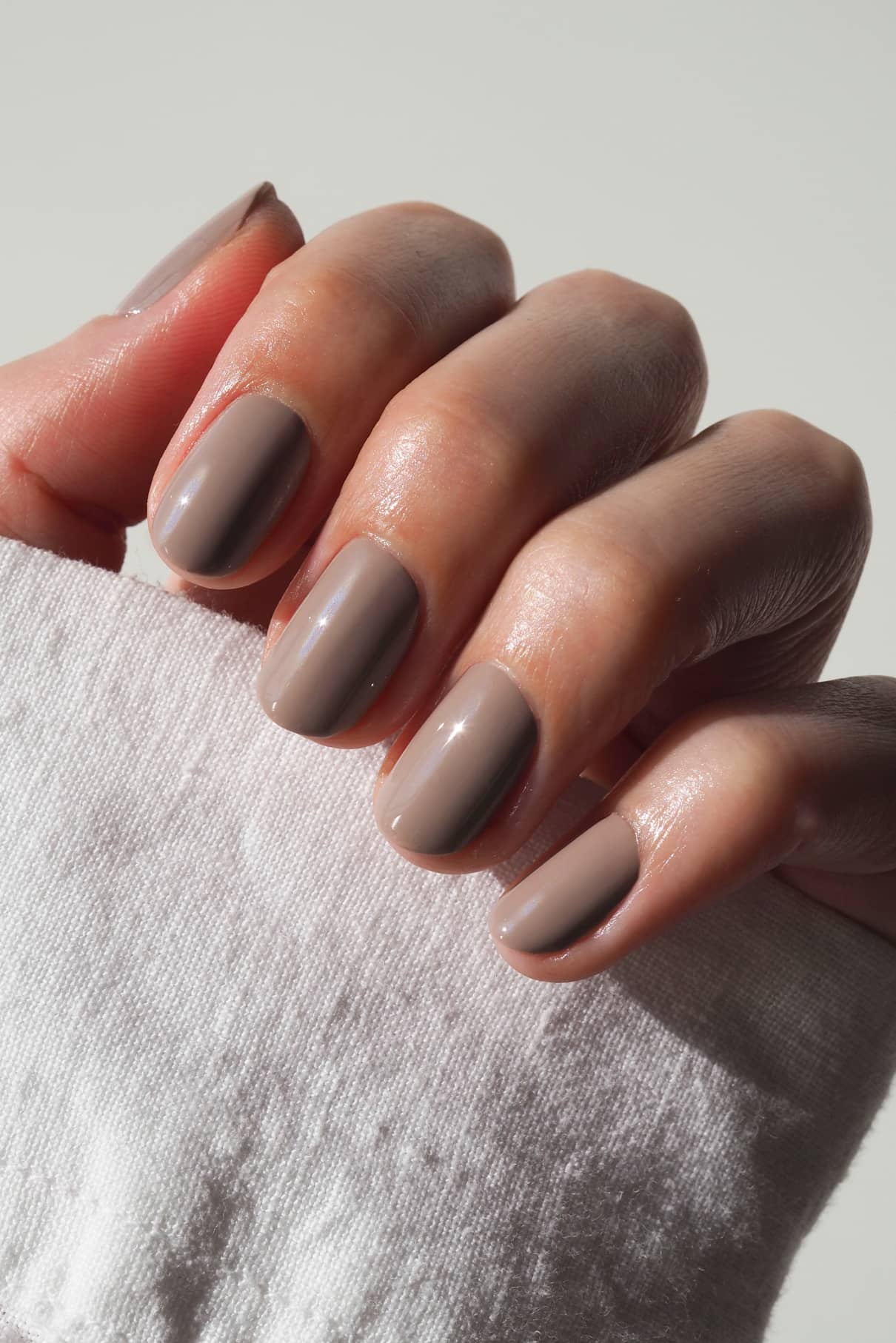 Eine Hand mit kurzen, sauber manikürten Nägeln, die in glänzendem Gitti Nr. 019 (taupe mit beigen Untertönen) auf einem weißen, strukturierten Stoff ruht.