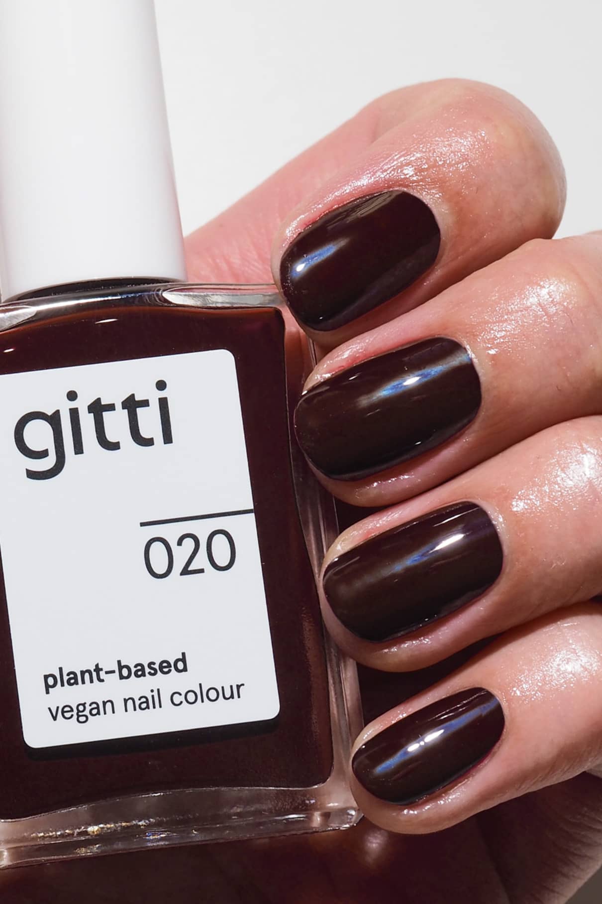 Eine Hand mit kurzen, sauber manikürten und in tiefem Rot lackierten Nägeln hält eine Flasche gitti no. 020 vegane Nagelfarbe auf Pflanzenbasis.