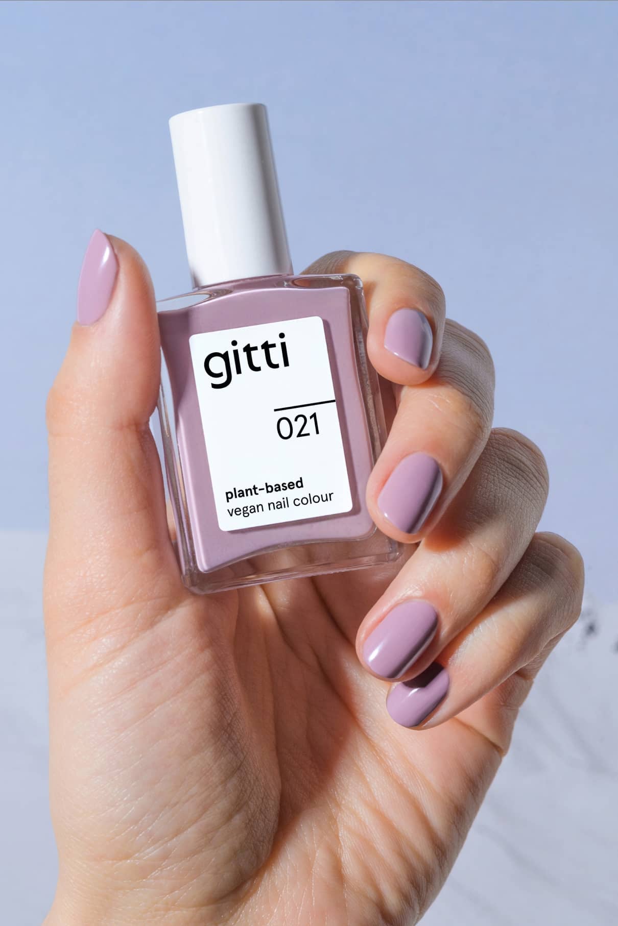 Eine Hand mit lavendelfarbenen Nägeln in einem pudrigen Lila-Ton hält eine Flasche gitti no. 021 pflanzlicher veganer Nagelfarbe vor einem hellen Hintergrund.