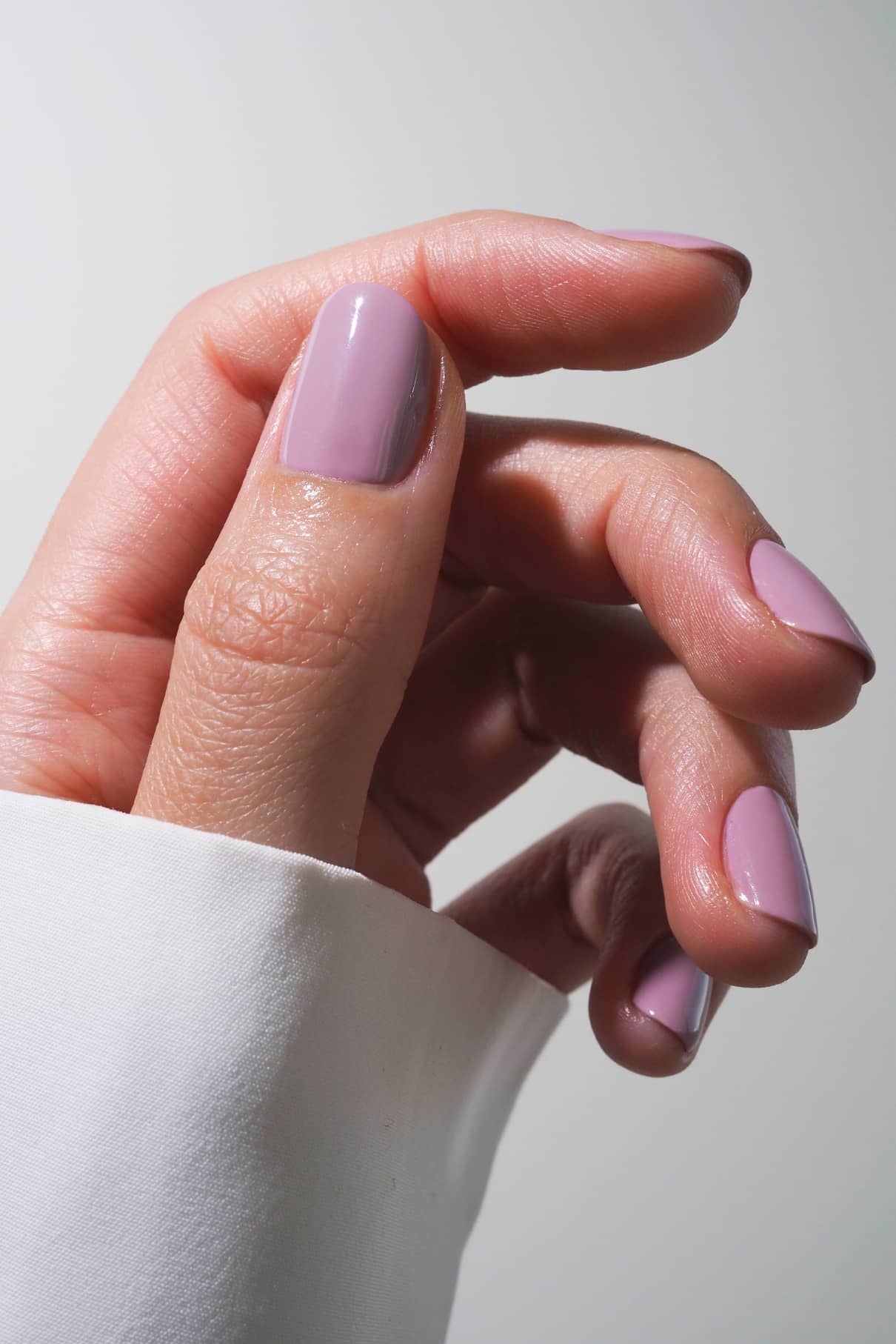 Eine Hand mit kurzen, sauber manikürten Nägeln, lackiert in gitti no. 021, einem pudrigen Fliederton, vor einem neutralen Hintergrund, auf dem ein Teil eines weißen Ärmels zu sehen ist.