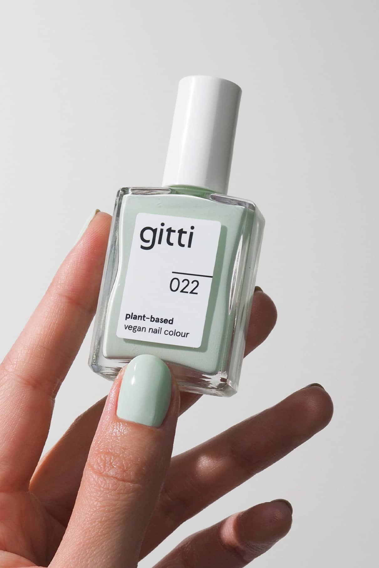 Eine Hand mit in frischem Mintgrün lackierten Nägeln hält eine Flasche gitti no. 022 pflanzlicher veganer Nagelfarbe vor einem schlichten Hintergrund.