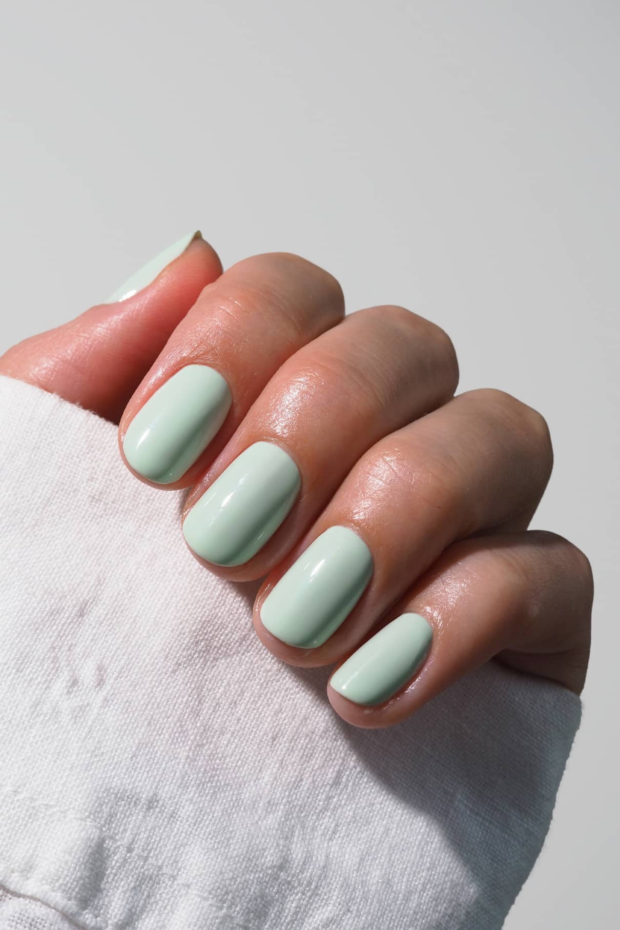Eine Hand mit kurzen, sauber manikürten Nägeln, lackiert in gitti no. 022, einem frischen mintgrünen Farbton, teilweise bedeckt von einem weißen Ärmel.