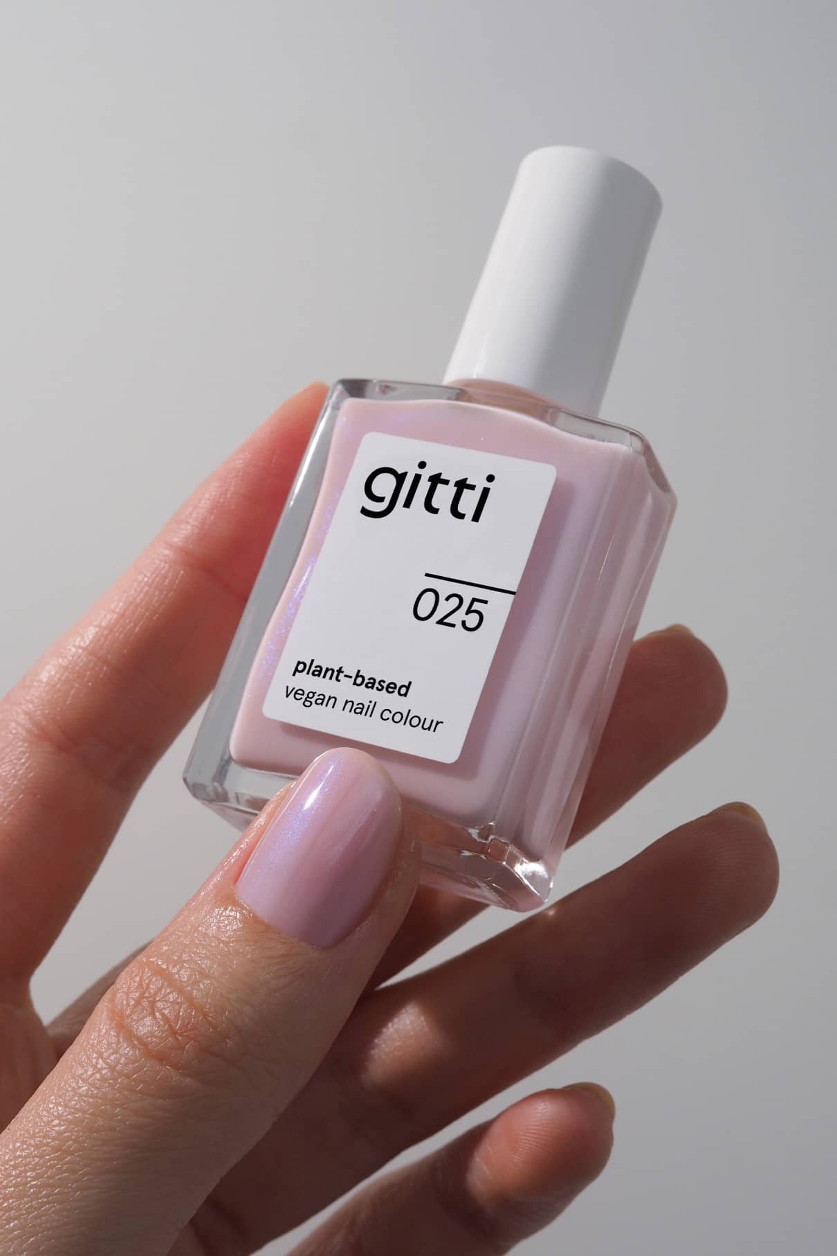 Eine Hand hält eine Flasche mit gitti no. 025 pflanzliche vegane Nagelfarbe; die Nägel sind in demselben halbtransparenten, hellrosa Farbton mit bläulichem Schimmer lackiert.