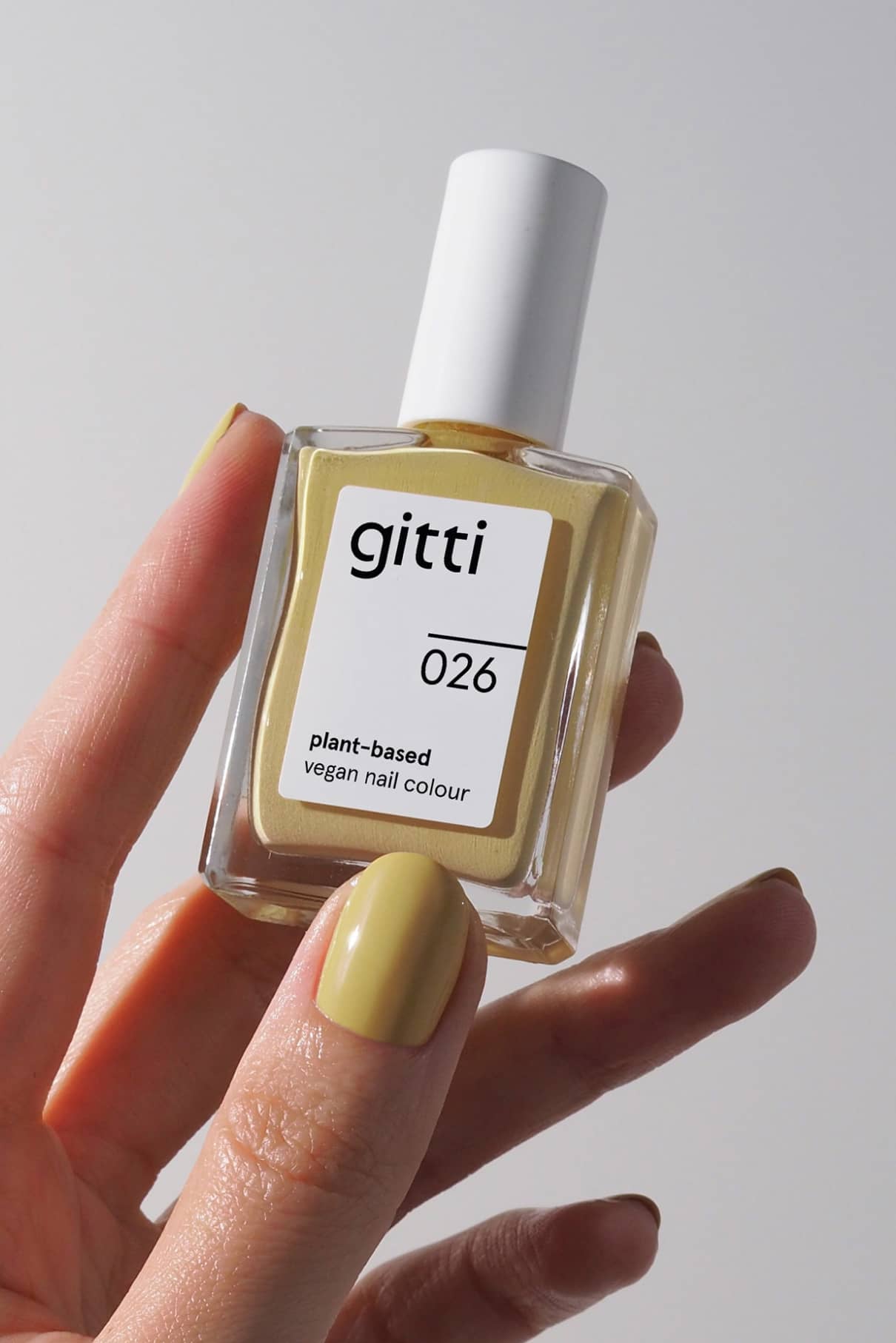 Eine Hand mit gelb lackierten Nägeln hält eine Flasche von gitti no. 026, eine pflanzliche, vegane Nagelfarbe in Pastellgelb mit grünem Touch, vor einem schlichten Hintergrund.