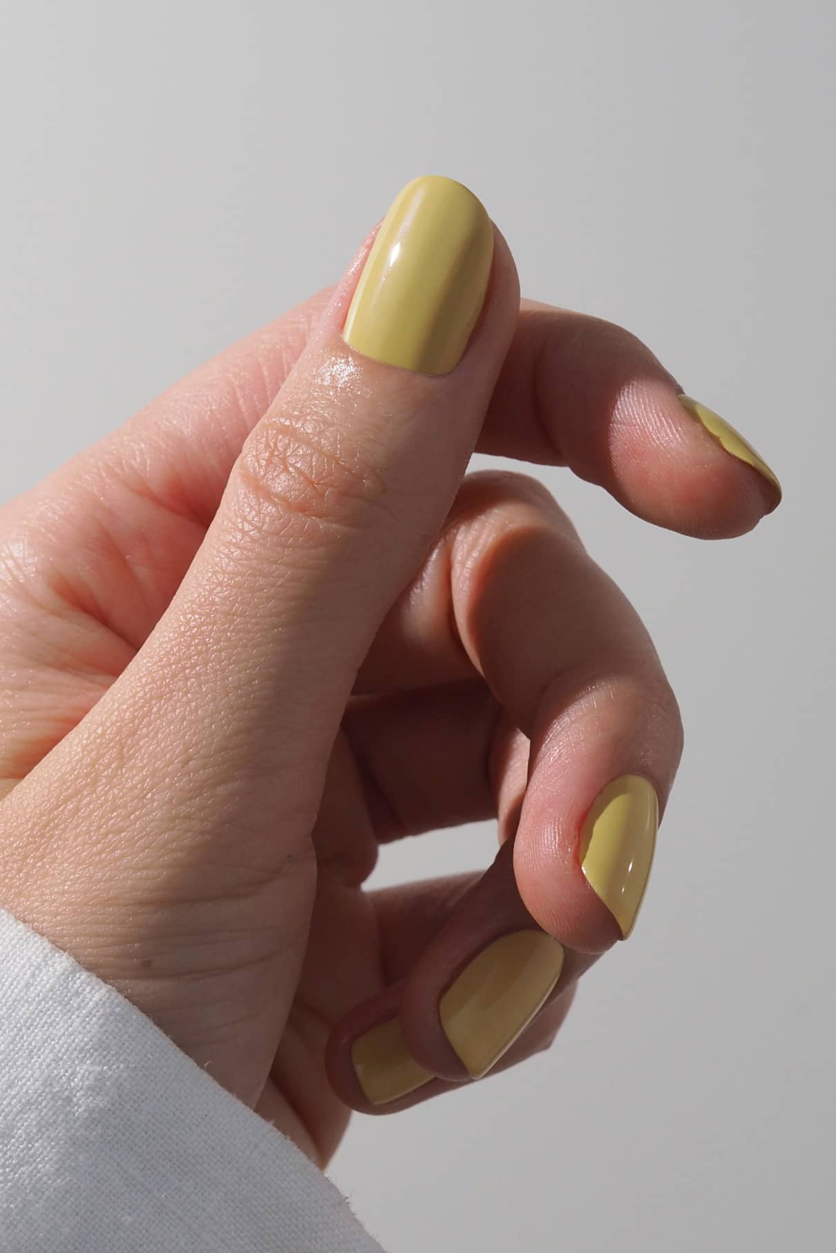 Eine Hand mit kurzen, sauber manikürten Nägeln, lackiert in glänzendem Gitti Nr. 026, einem Pastellgelb mit grünem Touch, vor einem unifarbenen Hintergrund abgebildet.