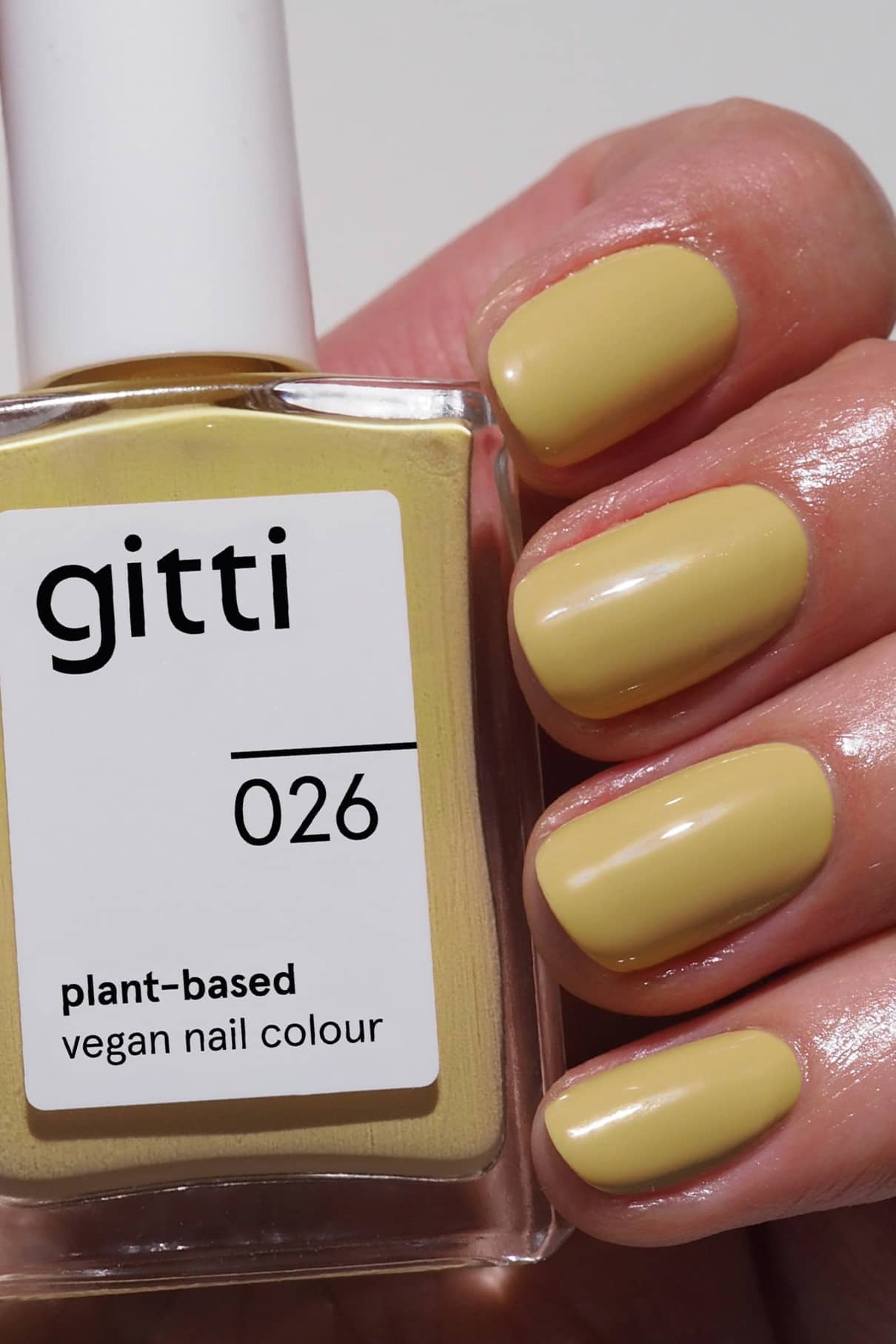 Eine Hand mit kurzen Nägeln in pastelligem Gelb hält eine Flasche gitti no. 026, einen veganen, pflanzenbasierten Nagellack im selben Pastellton - für einen natürlichen Look mit grünem Touch.