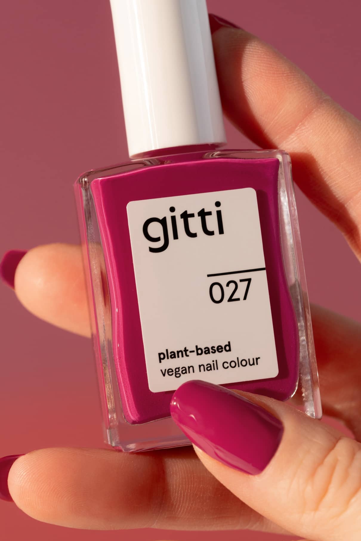 Eine Hand mit leuchtend magentafarbenem Nagellack hält eine Flasche gitti no. 027 pflanzlicher veganer Nagellack in Fuchsia-Pink vor einem rosa Hintergrund.