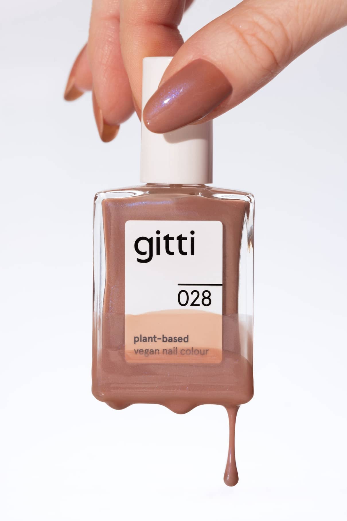 Eine Hand mit lackierten Nägeln hält eine Flasche gitti no. 028 pflanzlicher veganer Nagellack in warmem Mokkabraun, mit dezentem blauen Schimmer und Lack, der aus der Flasche tropft.