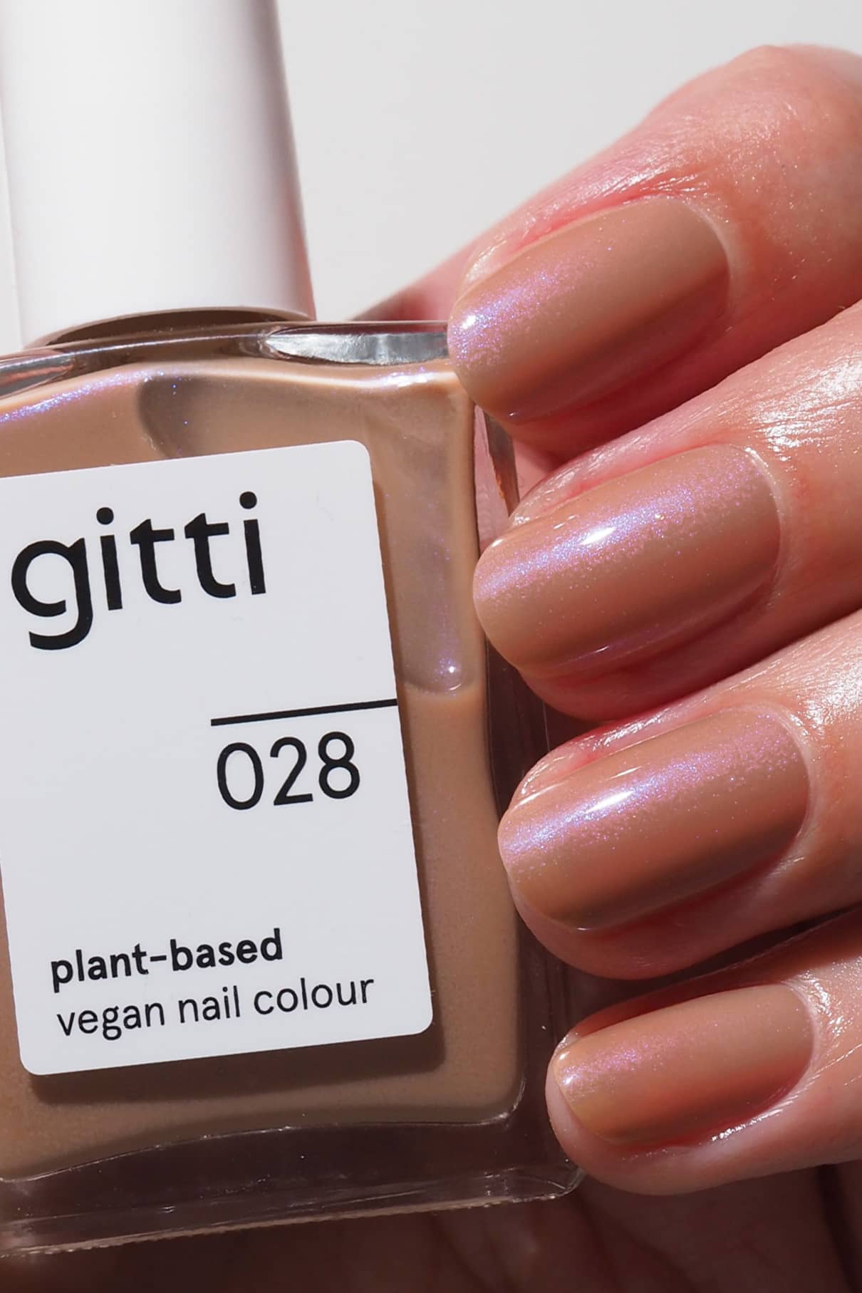 Eine Hand mit kurzen, manikürten Nägeln, die in warmem mokkabraunem Nude-Lack mit blauem Schimmer lackiert sind, hält eine Flasche mit der Aufschrift gitti no. 028 vegane Nagelfarbe auf Pflanzenbasis.