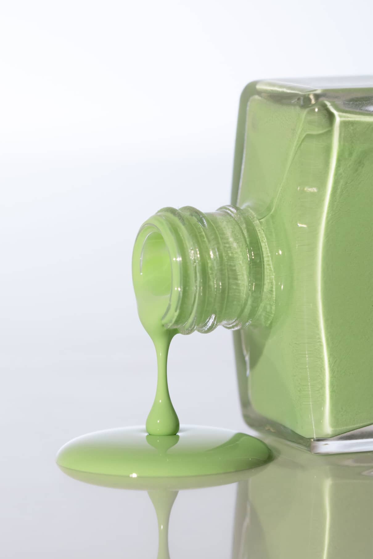 Eine quadratische Glasflasche mit gitti no. 029 Nagellack ist umgekippt, der frische, belebende Grünton läuft aus und sammelt sich auf einer spiegelnden Oberfläche.