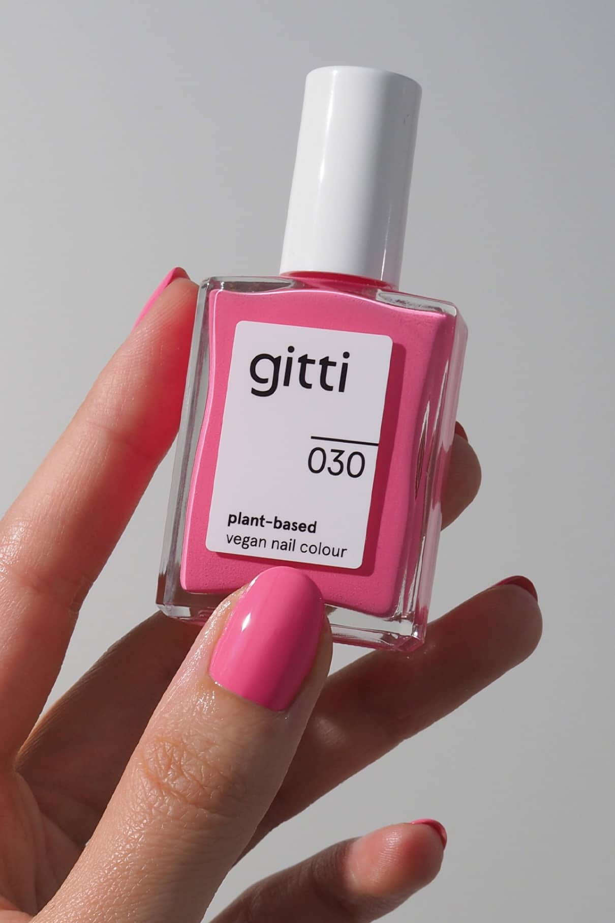 Eine Hand mit rosa lackierten Nägeln hält eine quadratische Flasche von gitti no. 030, eine pflanzliche, vegane Nagelfarbe in leuchtendem Pink, vor einem schlichten Hintergrund.