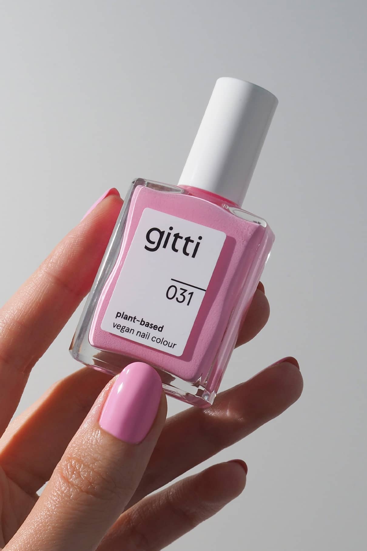 Eine Hand mit rosa lackierten Nägeln hält eine Flasche gitti no. 031, eine pflanzliche, vegane Nagelfarbe in verträumtem Pastellrosa, die schicke Retro-Vibes versprüht.