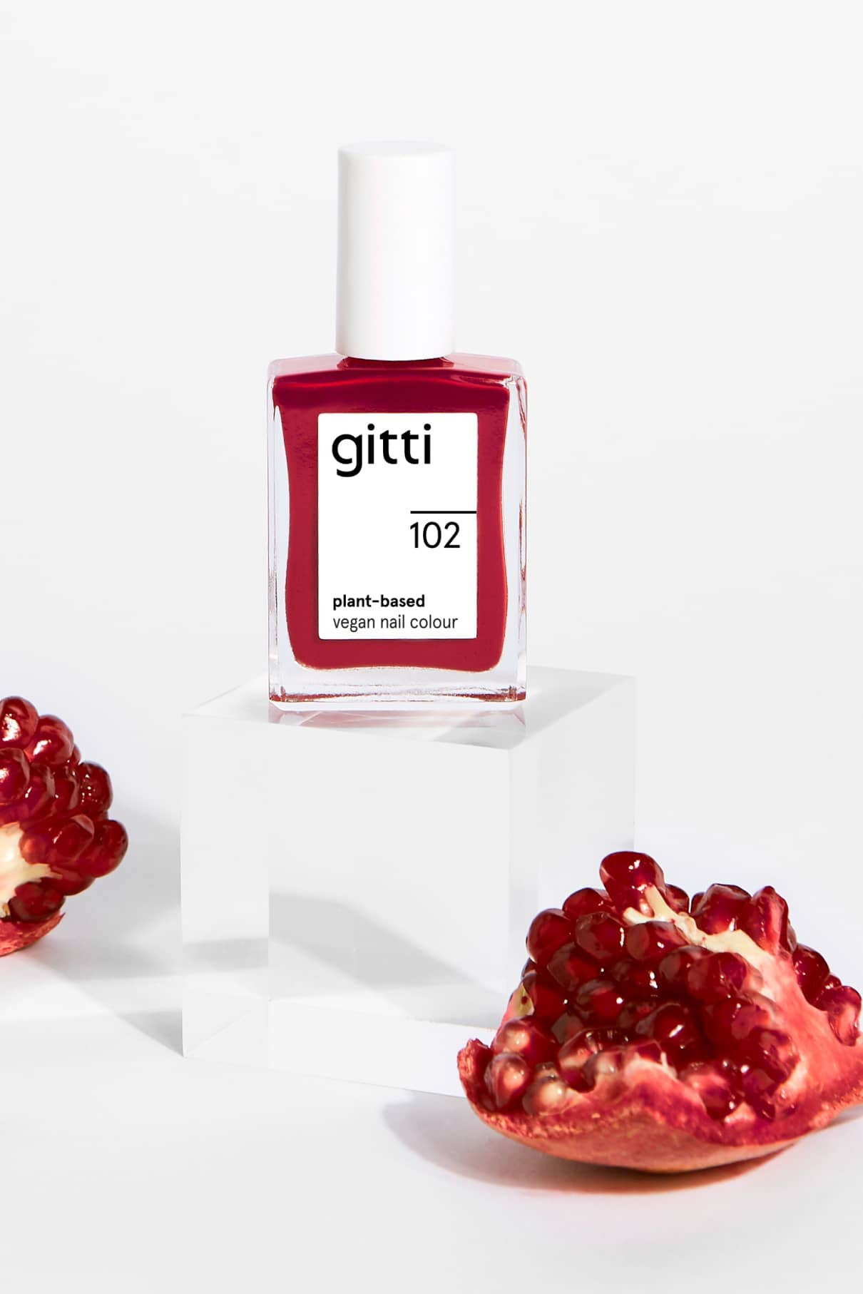 Eine Flasche gitti no. 102 pflanzlicher, veganer Nagellack, ein von Rotwein inspirierter Farbton, wird auf einem durchsichtigen Block mit Granatapfelstücken auf weißem Hintergrund präsentiert.