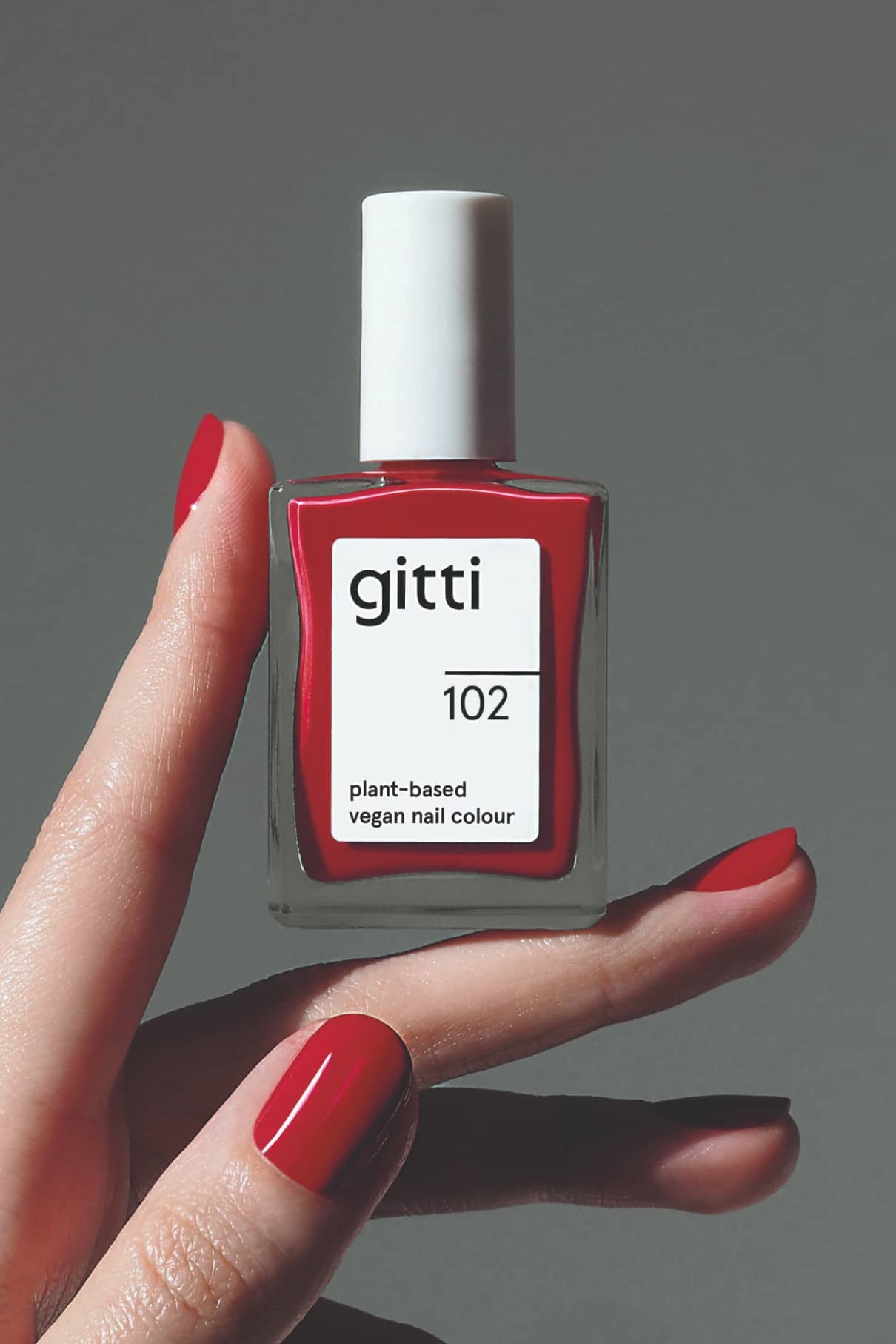 Eine Hand mit rot lackierten Nägeln hält eine Flasche von gitti no. 102, eine pflanzliche, vegane Nagelfarbe in einem von Rotwein inspirierten Farbton, vor einem schlichten grauen Hintergrund.