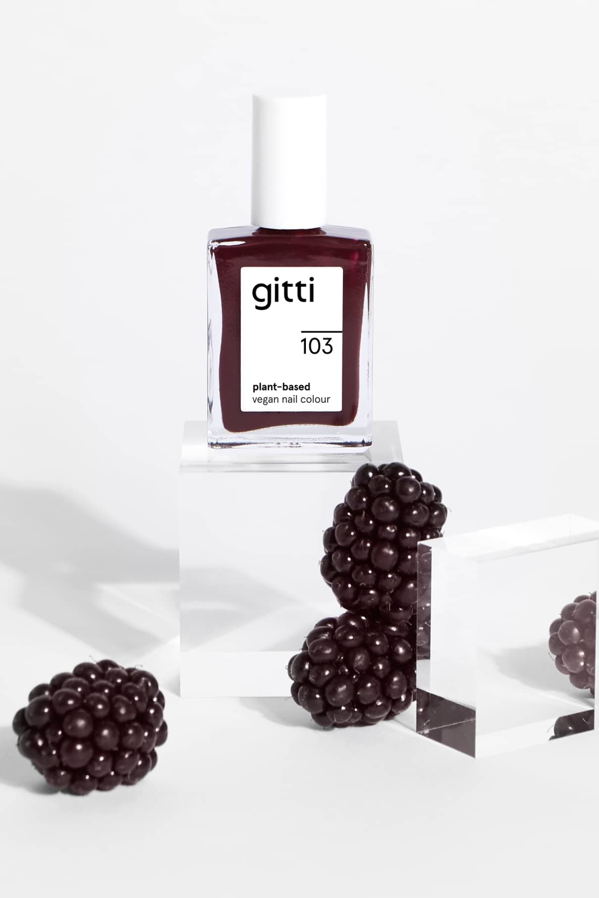 Eine Flasche gitti no. 103 pflanzliche vegane Nagelfarbe in dunklem Aubergine ist auf klaren Acrylblöcken ausgestellt, umgeben von Brombeeren.