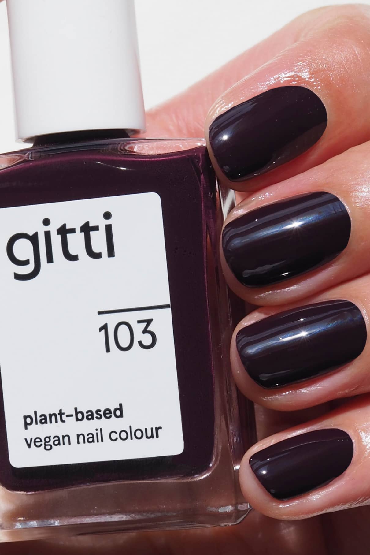 Eine Hand mit kurzen, sauber manikürten Nägeln, die in einem dunklen Aubergine-Ton lackiert sind, hält eine Flasche gitti no. 103 pflanzlicher veganer Nagellack.