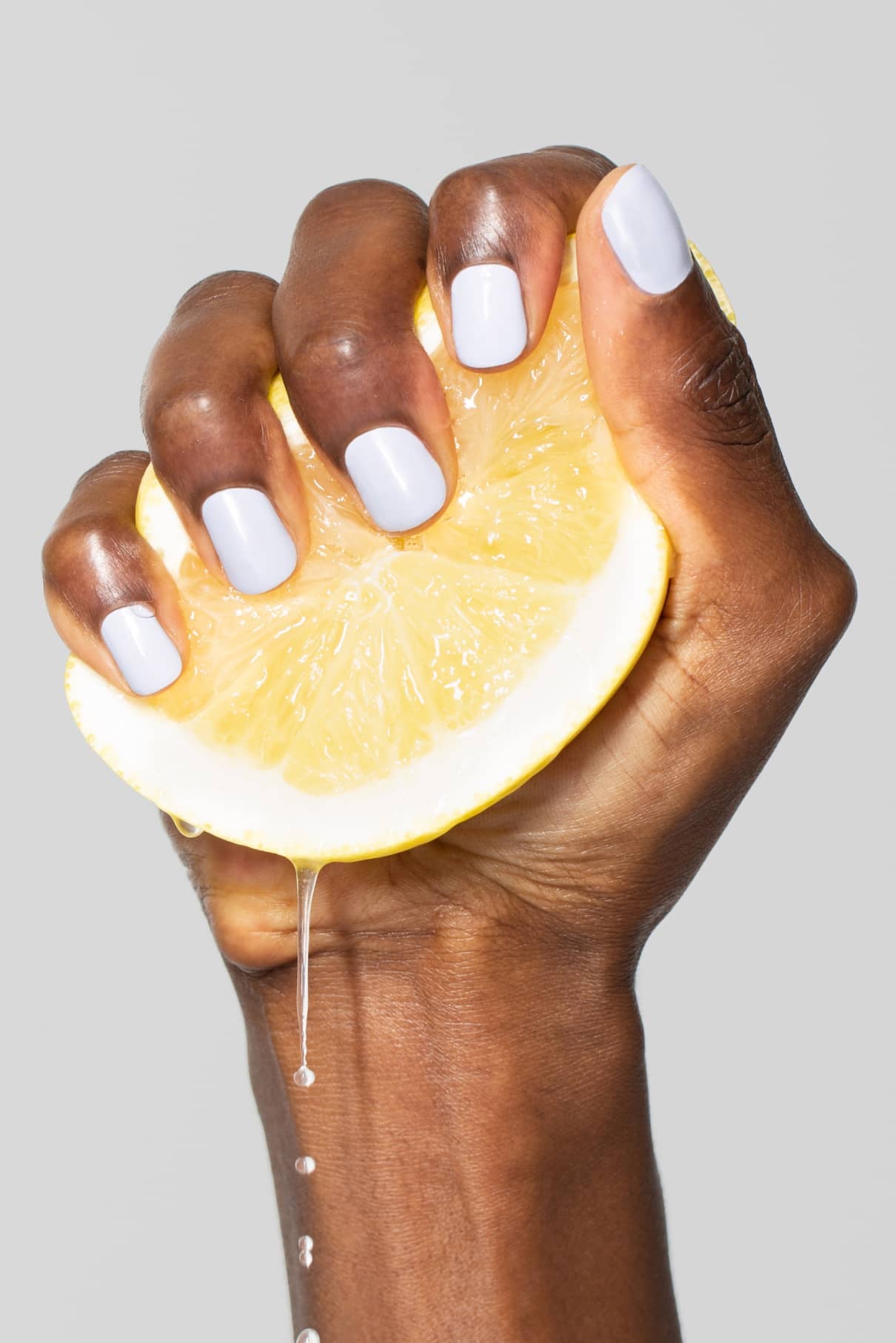 Eine Hand mit gitti no. 106 Nagellack in Pastellblau presst eine Zitronenscheibe aus, die Saft abgibt, vor einem schlichten grauen Hintergrund.