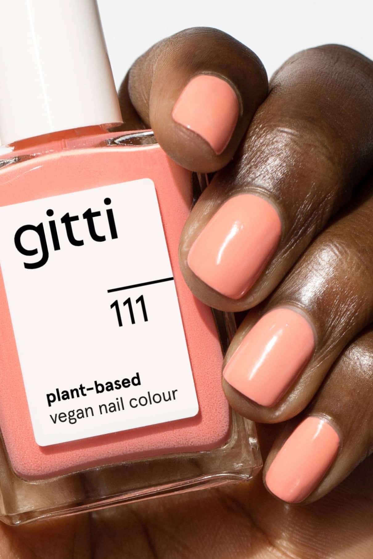 Eine Hand mit kurzen, sauber manikürten und in zartem Pfirsichrosa mit warmem Unterton lackierten Nägeln hält eine Flasche gitti no. 111 veganen Nagellack auf Pflanzenbasis.