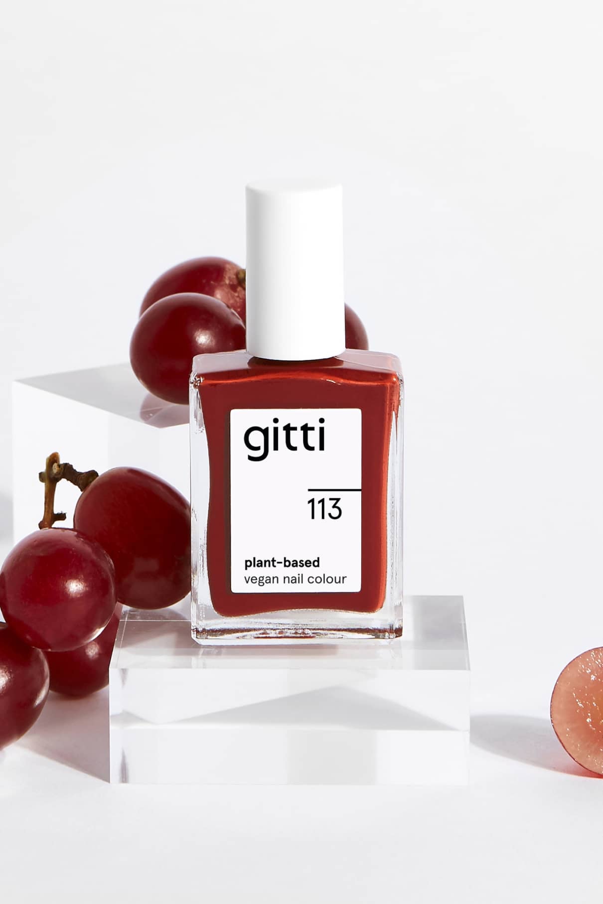 Eine Flasche gitti no. 113 pflanzlicher veganer Nagellack in braun-roter Farbe ist mit roten Trauben auf und neben durchsichtigen Acrylblöcken vor einem weißen Hintergrund abgebildet.
