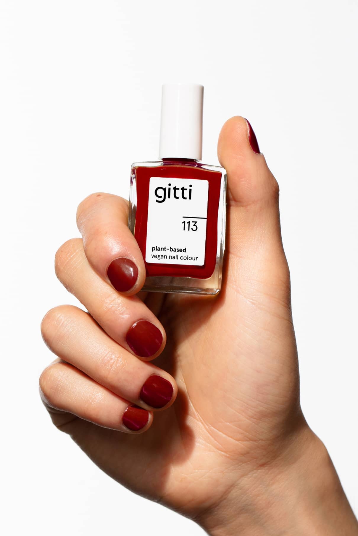 Eine Hand mit dunkelrot lackierten Nägeln hält eine Flasche mit gitti no. 113, einem veganen Nagellack auf pflanzlicher Basis, und hebt diesen warmen Farbton vor einem weißen Hintergrund hervor.