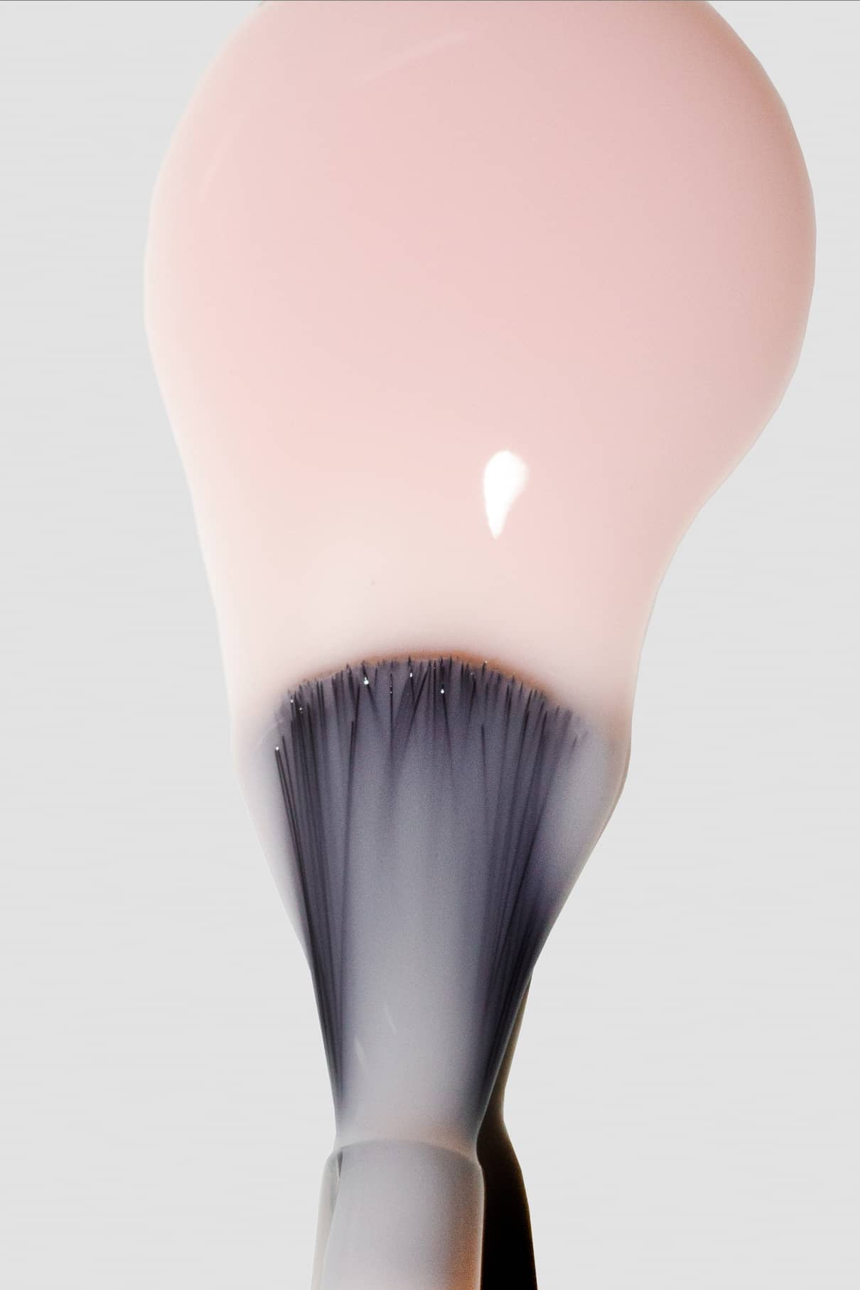Nahaufnahme eines Make-up-Pinsels mit halbtransparenter, blassrosa gitti no. 114 Flüssigfoundation, die sich auf den Borsten vor einem einfarbigen Hintergrund sammelt.