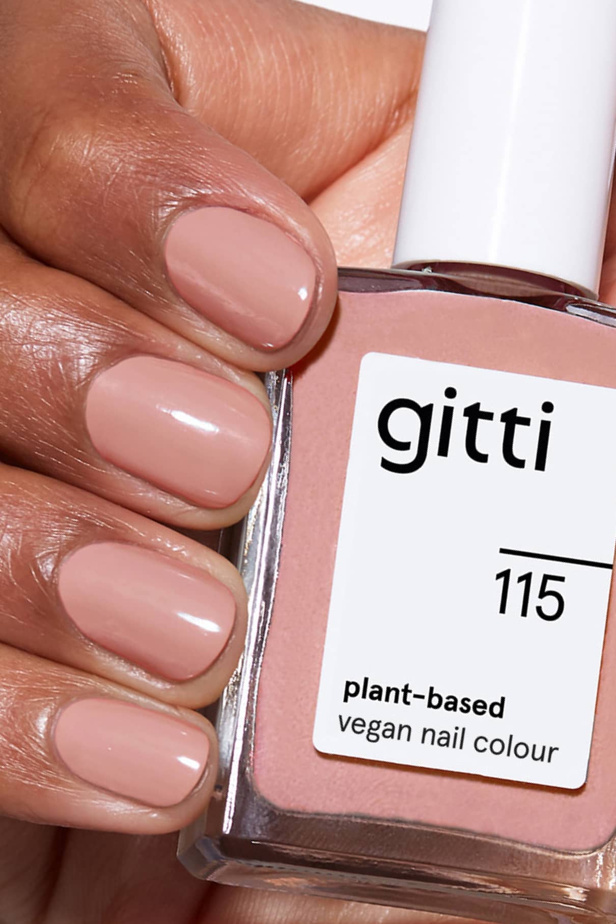 Eine Hand mit kurzen, sauber manikürten Nägeln, die in einem nude-rosa-beigen Farbton lackiert sind, hält eine Flasche gitti no. 115 veganen Nagellack auf Pflanzenbasis.