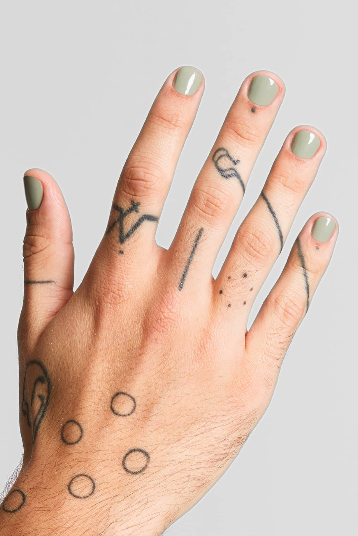 Eine linke Hand mit Nägeln, die in Gitti Nr. 121, einem sanften, gedeckten Grünton, und mehreren schwarzen Strich- und Punkttattoos an den Fingern und auf dem Handrücken.