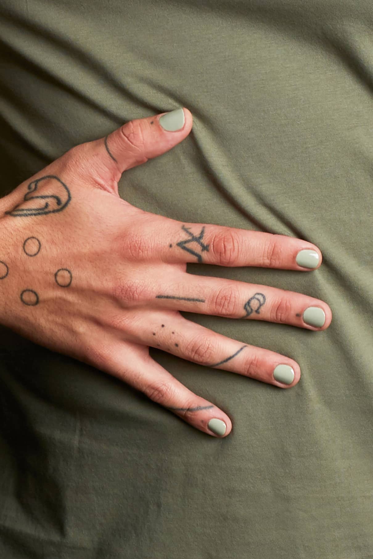 Eine Hand mit hellgrün lackierten Nägeln (gitti Nr. 121) und schwarzen Strich-Tattoos ruht auf einem olivgrünen Stoffhintergrund, wobei die Nagelfarbe an eine moderne salbeigrüne Wandfarbe erinnert.