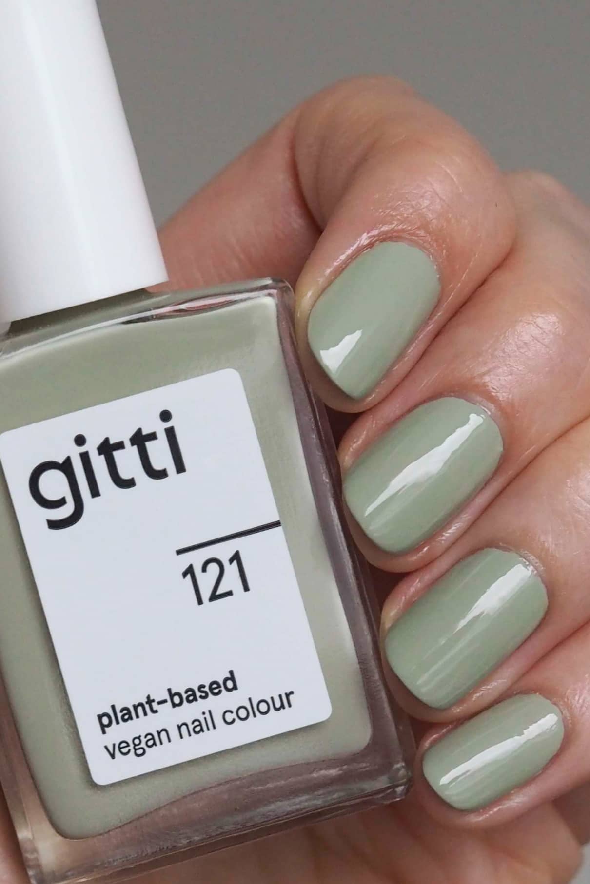 Eine Hand mit kurzen, sauber manikürten und in dezentem Grün lackierten Nägeln hält eine Flasche gitti no. 121, einem veganen Nagellack auf Pflanzenbasis.