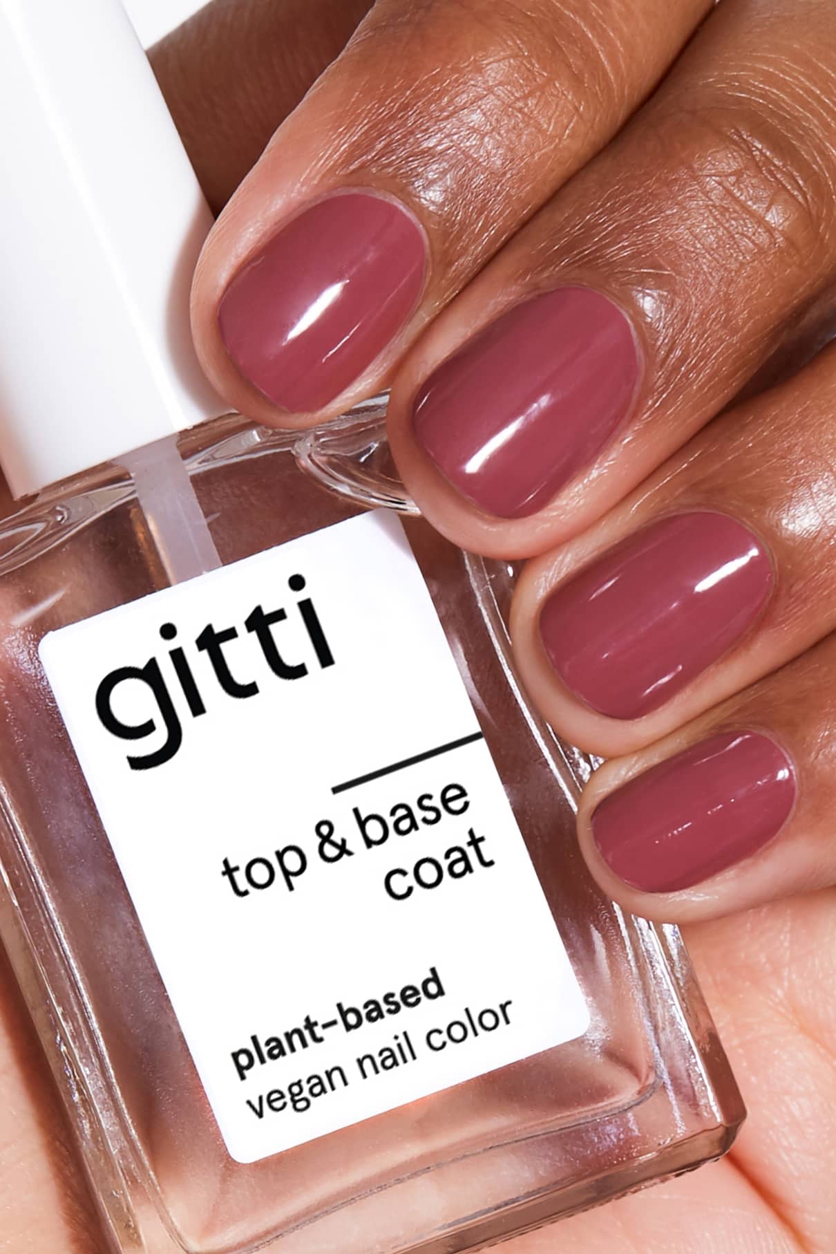 Eine Hand mit kurzen, sauber manikürten Nägeln, die in gitti no. 126 (ein sanftes Rot) lackiert sind, hält eine durchsichtige Flasche mit der Aufschrift gitti top & base coat, eine vegane Nagelfarbe auf Pflanzenbasis.