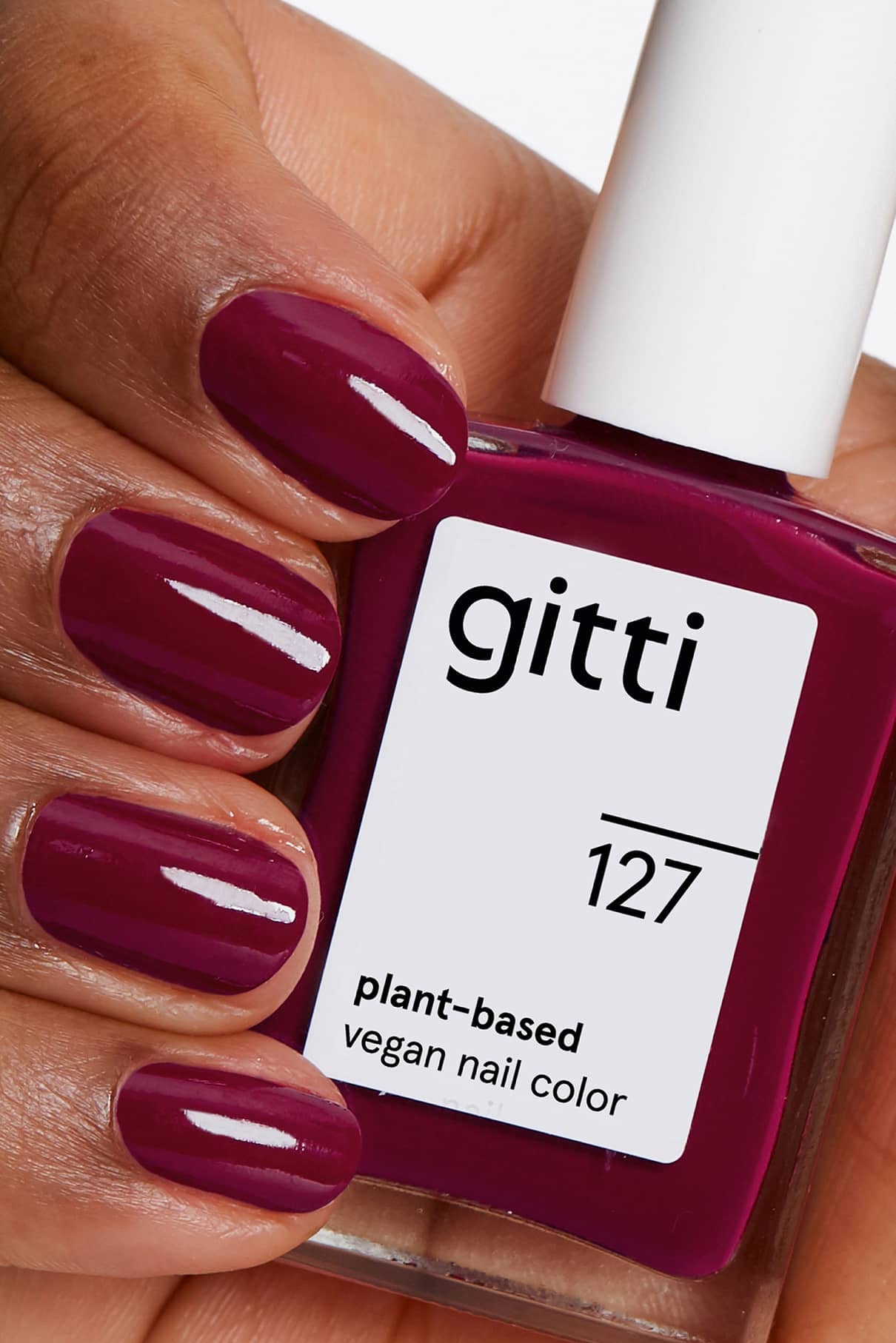 Eine Hand mit kurzen, sauber manikürten Nägeln, die in glänzendem dunkelrosa beeriger Pflaumenton lackiert sind, hält eine Flasche gitti no. 127 vegane Nagelfarbe auf Pflanzenbasis.