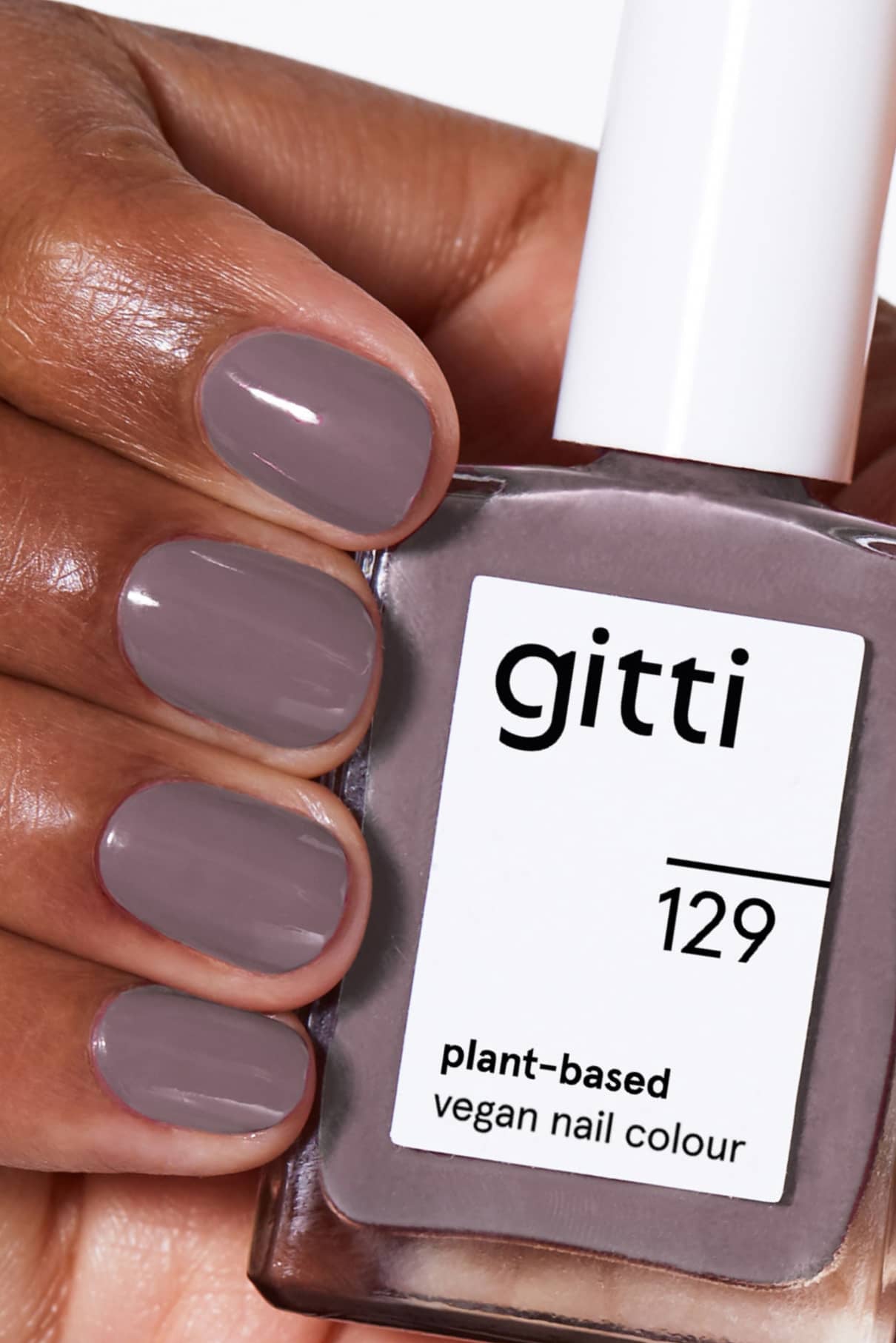 Eine Hand mit eleganten, taupefarbenen Nägeln hält ein Fläschchen von gitti no. 129, einer pflanzlichen, veganen Nagelfarbe in einem modernen Grau-Violett-Ton.