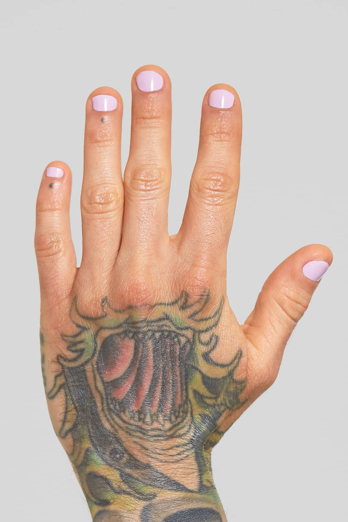 Eine tätowierte linke Hand mit pastelllila gitti no. 132 lackierten Nägeln ist vor einem schlichten grauen Hintergrund abgebildet.