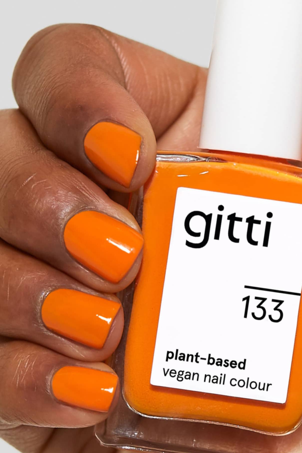Eine Hand mit orange lackierten Nägeln in warmem Orange hält eine Flasche gitti no. 133 vegane Nagelfarbe auf Pflanzenbasis.