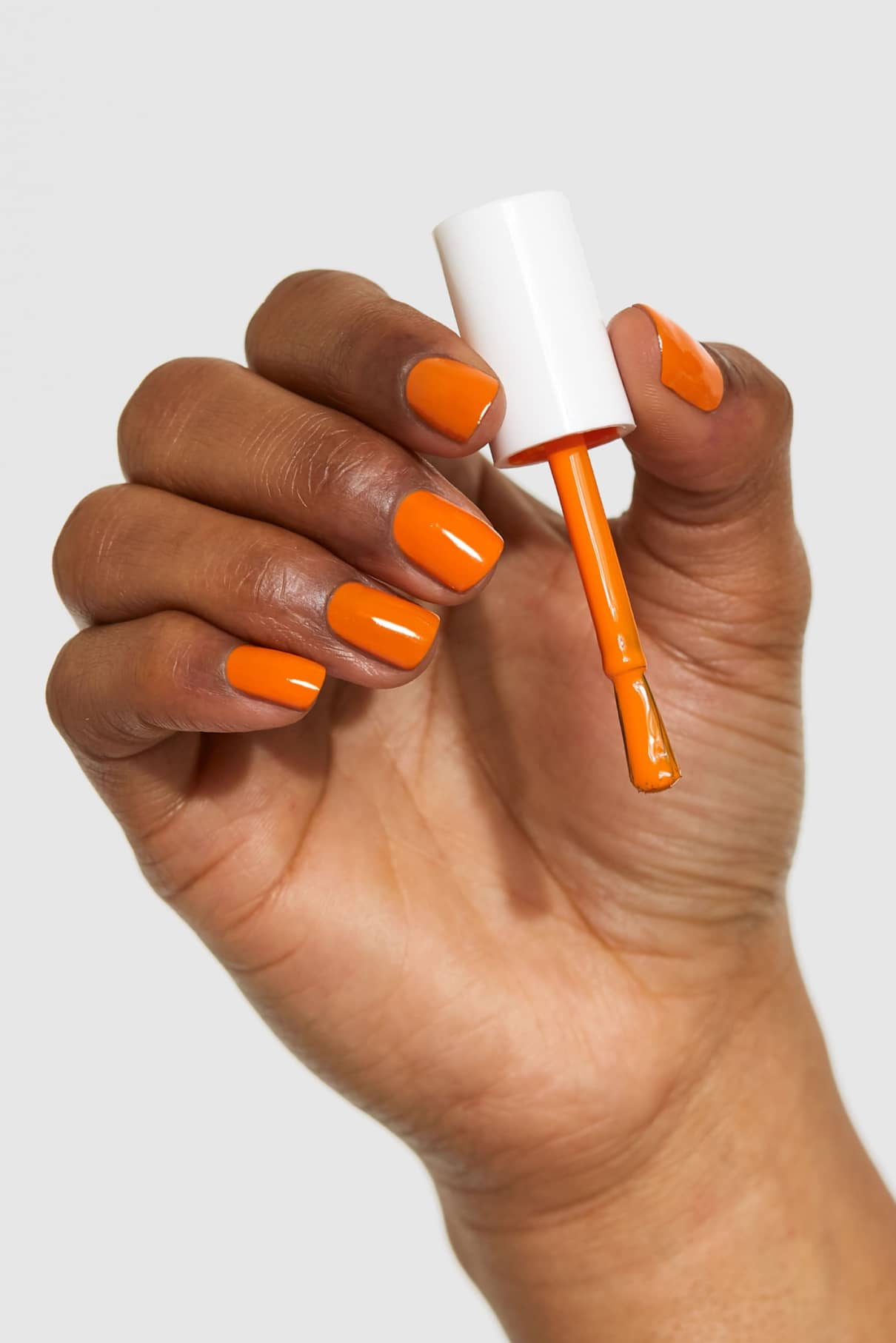 Eine Hand mit orange lackierten Nägeln hält eine Flasche gitti no. 133 Nagellack, Pinsel freigelegt, vor einem schlichten Hintergrund.
